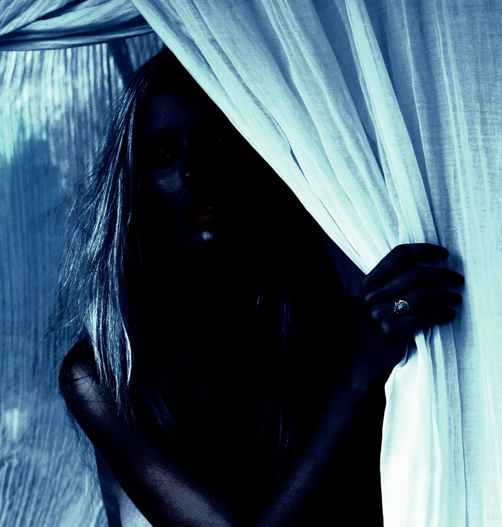 Woman in soft lighting with a mysterious expression, partially hidden behind curtains