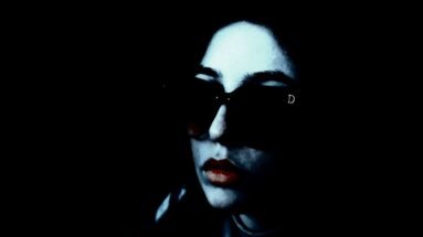 Woman sitting on a rooftop wearing sunglasses and a casual outfit, with a dark and moody atmosphere.