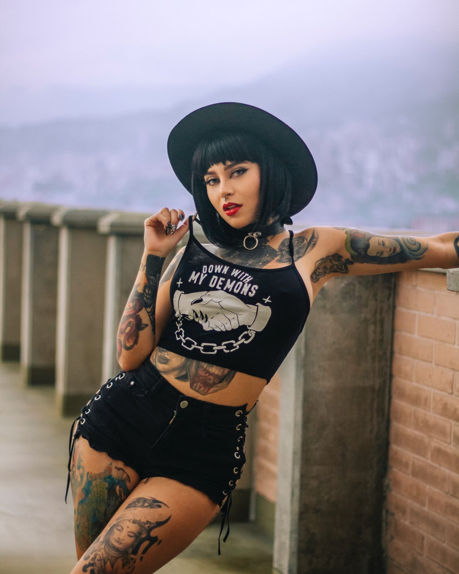 Tattooed goth woman in a black hat and shorts, leaning on a balcony with an urban background.
