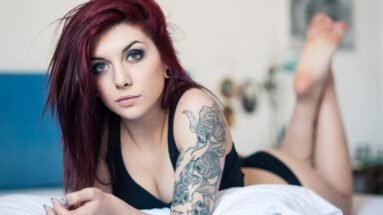 Young goth woman with red hair, lying on a bed and displaying her detailed arm tattoo.