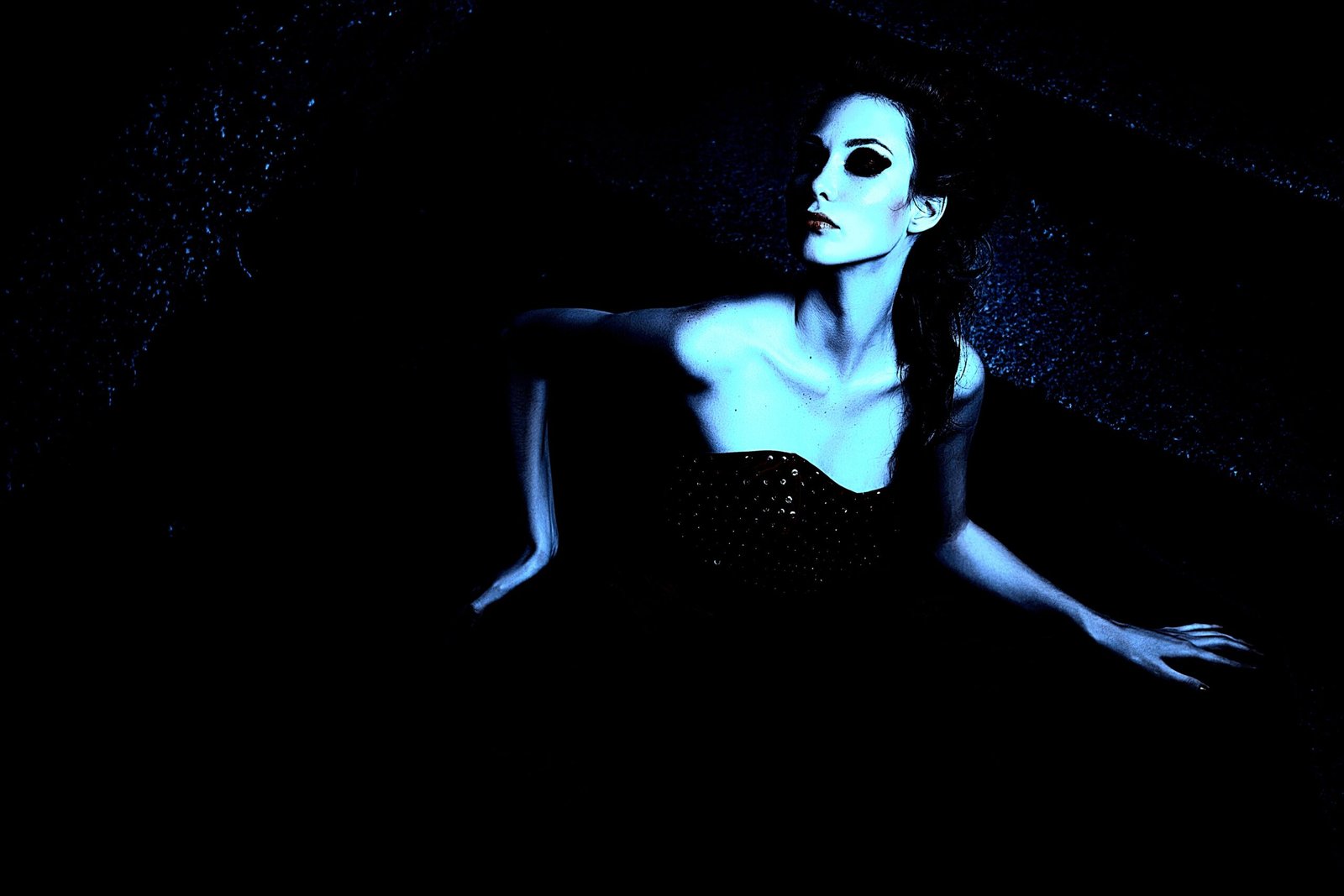 Gothic woman in a strapless black dress with dramatic makeup, lying on the ground in a dark, moody setting.