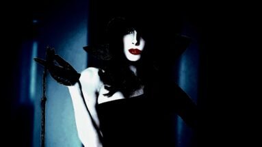 A woman with dark hair and dramatic makeup, dressed in a stylish, form-fitting outfit, exuding an air of sophistication and mystery.