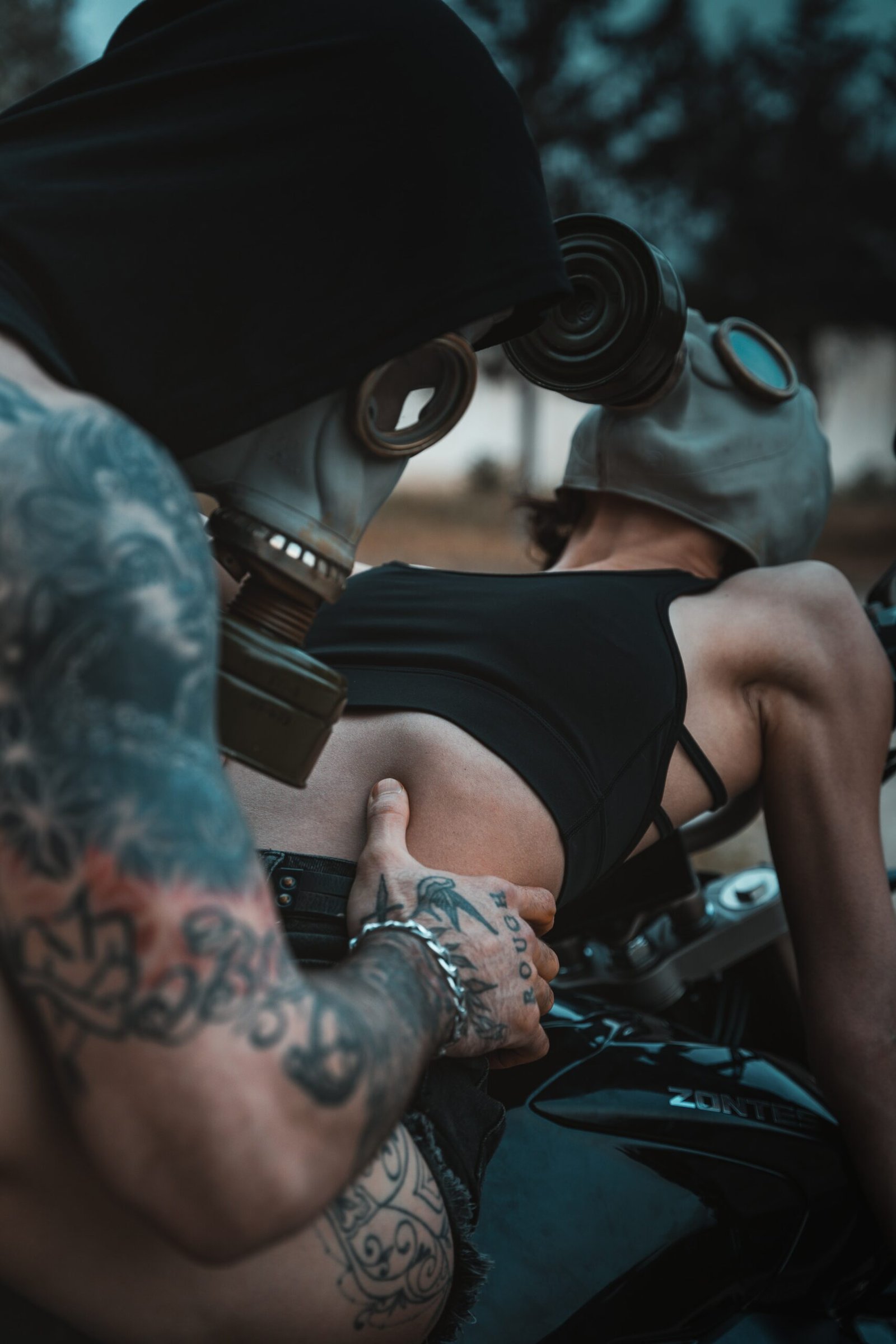 Two people wearing gas masks, with tattoos visible, riding a motorcycle in a post-apocalyptic style setting.
