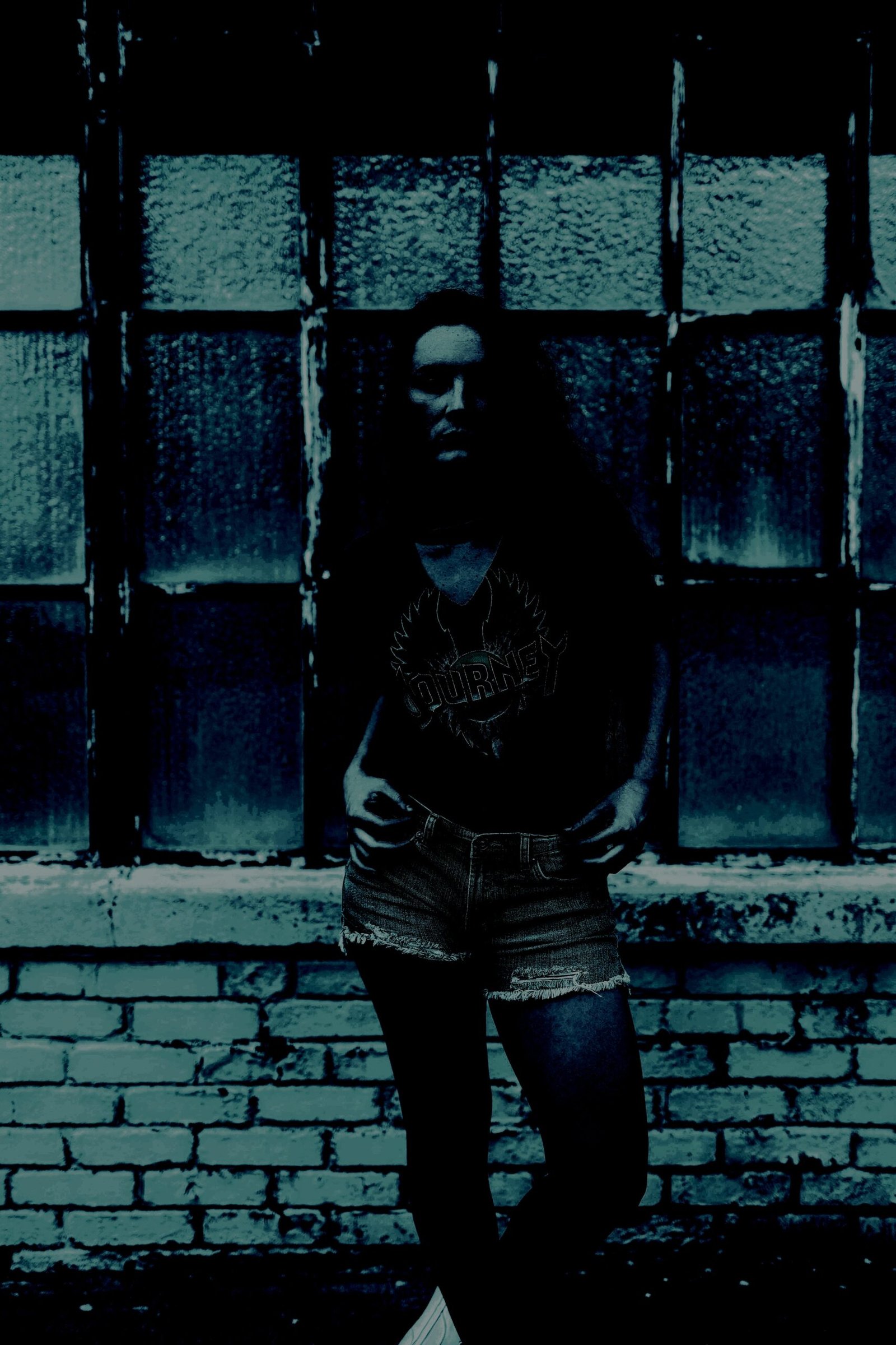 Woman in shorts and a Journey T-shirt standing against a grungy brick wall with a window.