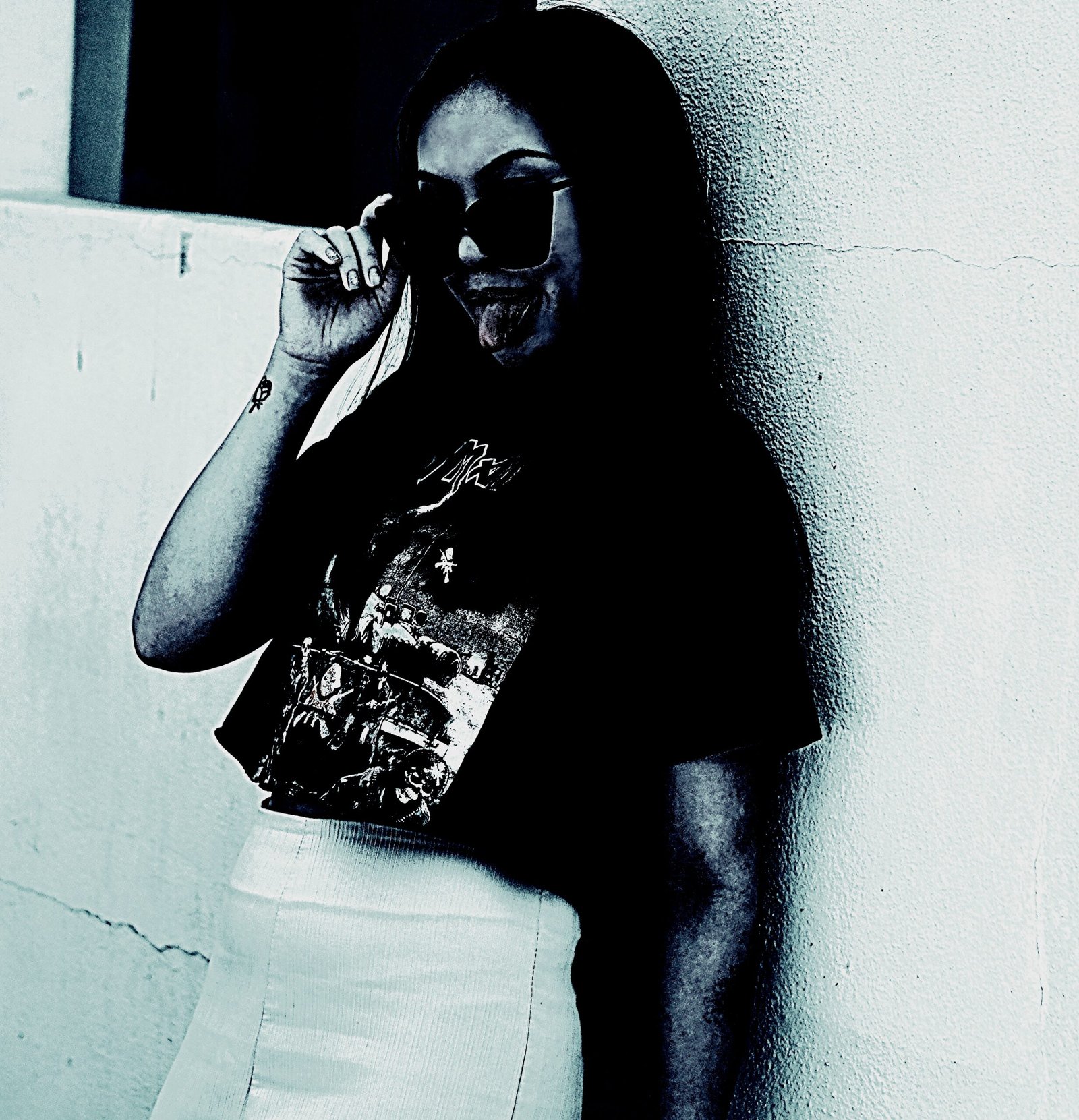 Woman with sunglasses posing in a dark setting wearing a graphic T-shirt and a light-colored skirt.