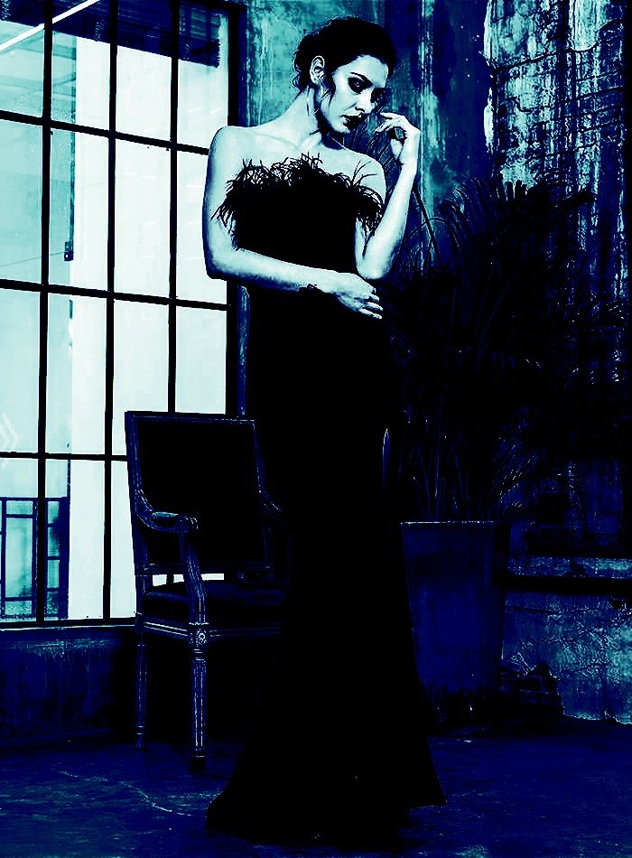 Woman in a gothic maxi dress with feather accents, posing in front of large windows