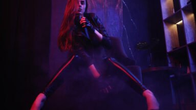 Gothic woman in a leather jacket and leggings, striking a confident pose in a dark, moody environment.