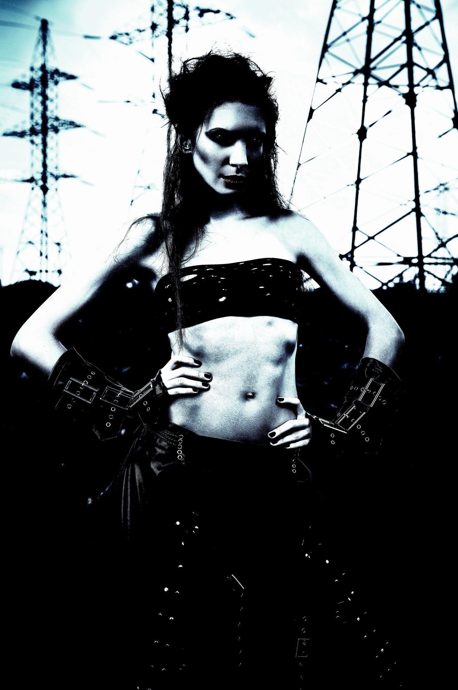 Gothic woman standing confidently in front of power lines, dressed in dark, alternative clothing with a fierce expression.