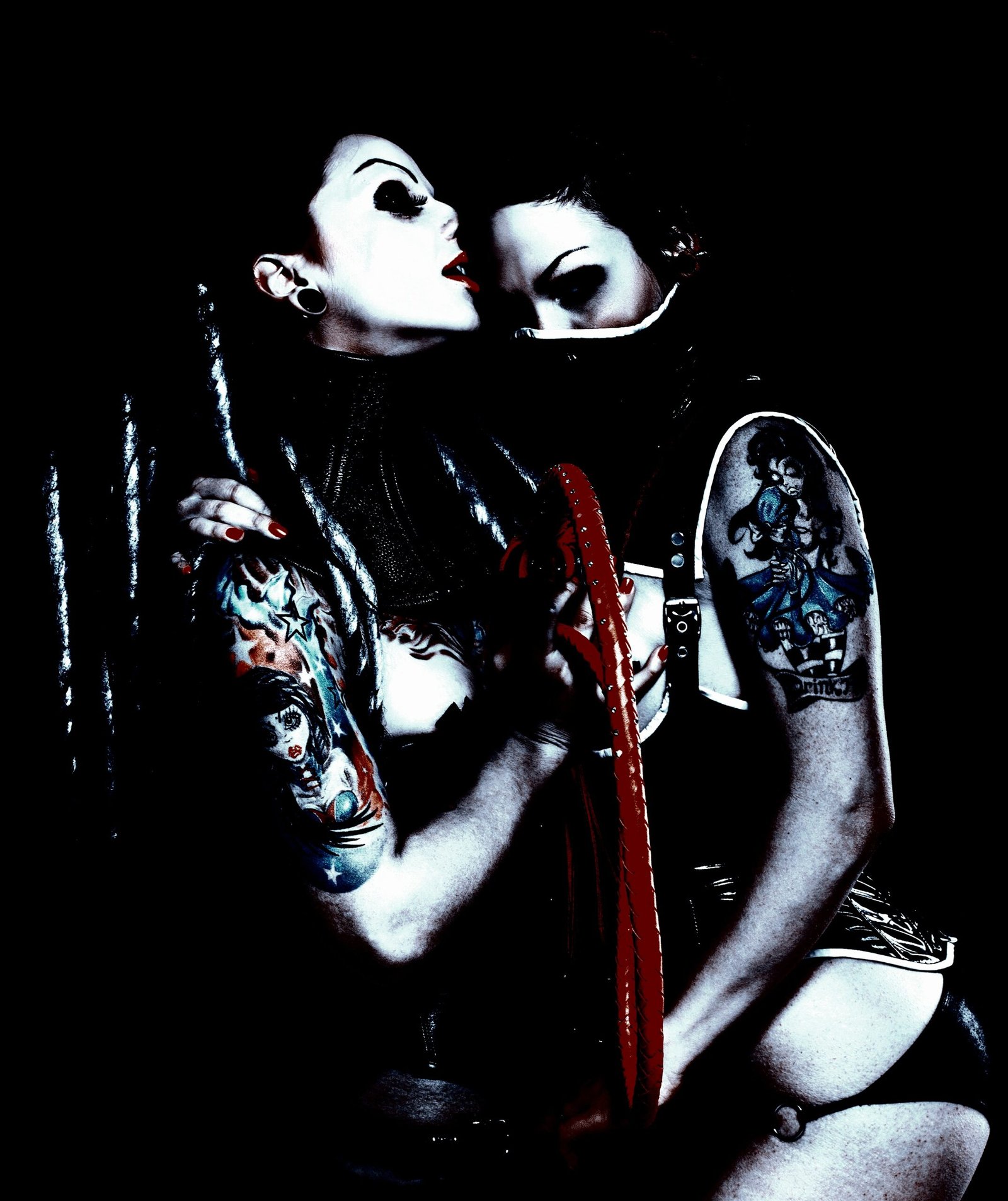 Two women with gothic tattoos, one wearing a mask and the other with bold makeup, in an intimate and darkly lit scene.