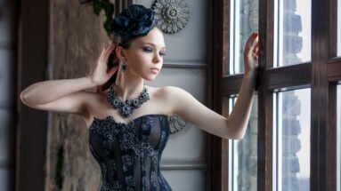 Woman in a black lace over bust corset looking out of a window with ornate decor, wearing a matching black fascinator.