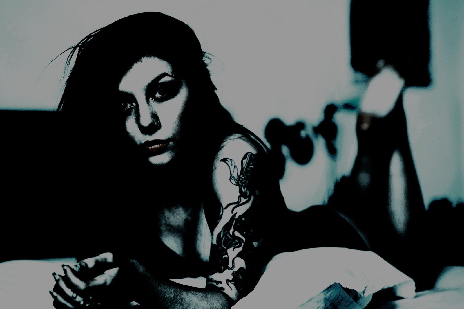 Woman with long dark hair and a gothic tattoo on her arm lying on a bed in a dark, moody setting.