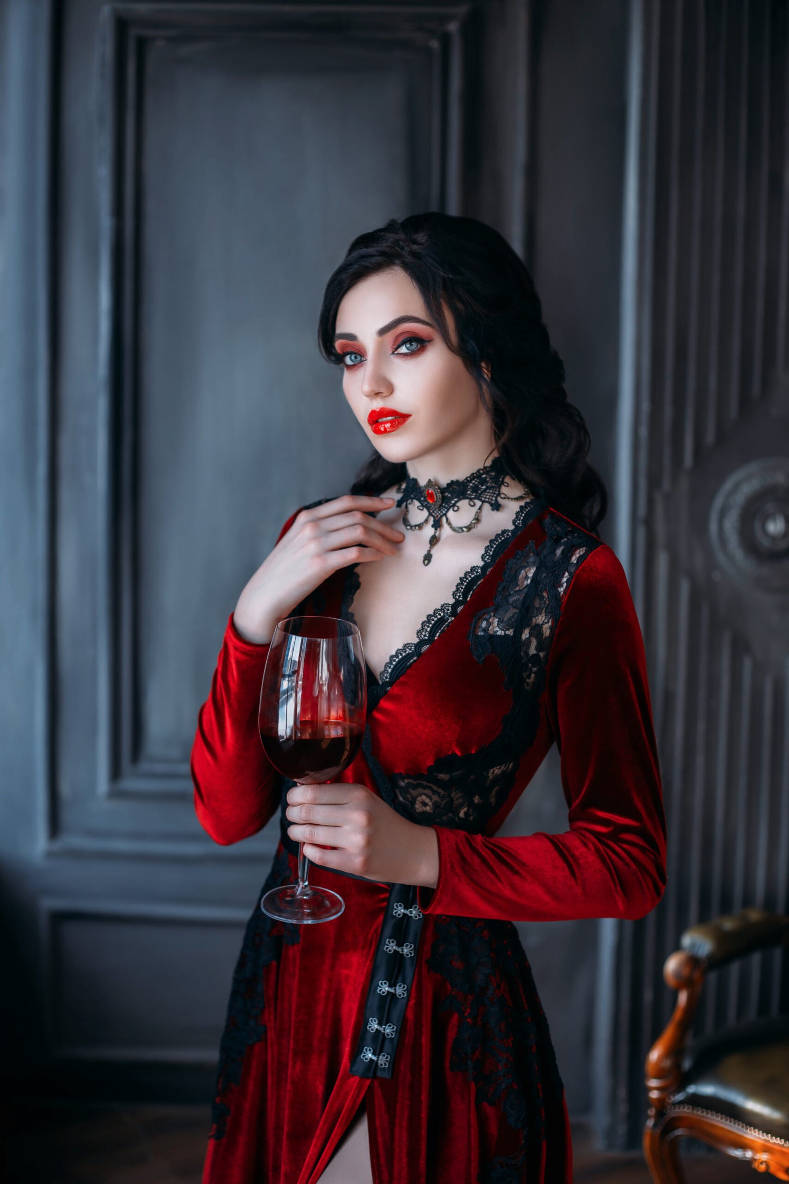 Woman in a deep red velvet gothic dress with black lace details, holding a glass of wine in a luxurious, dark interior.