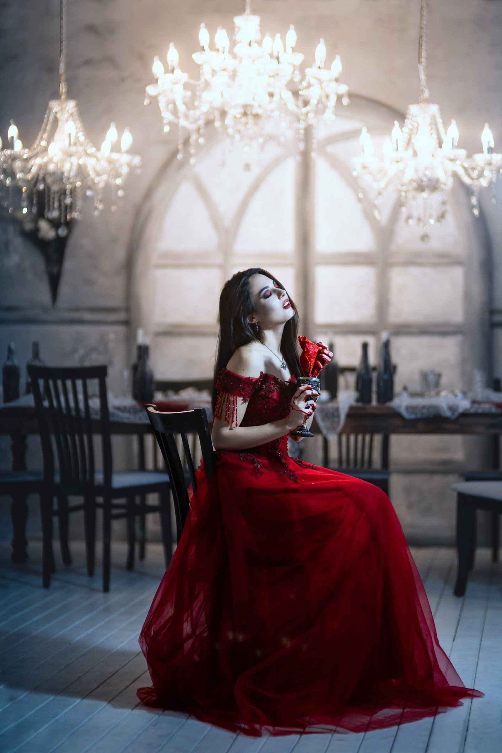 Gothic woman in a flowing red gown under dramatic lighting, seated in a dimly lit room with chandeliers.