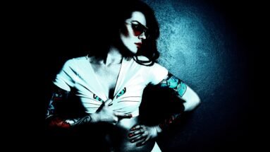 Woman with gothic tattoos on both arms pulling down her white top, with dramatic lighting and dark background.
