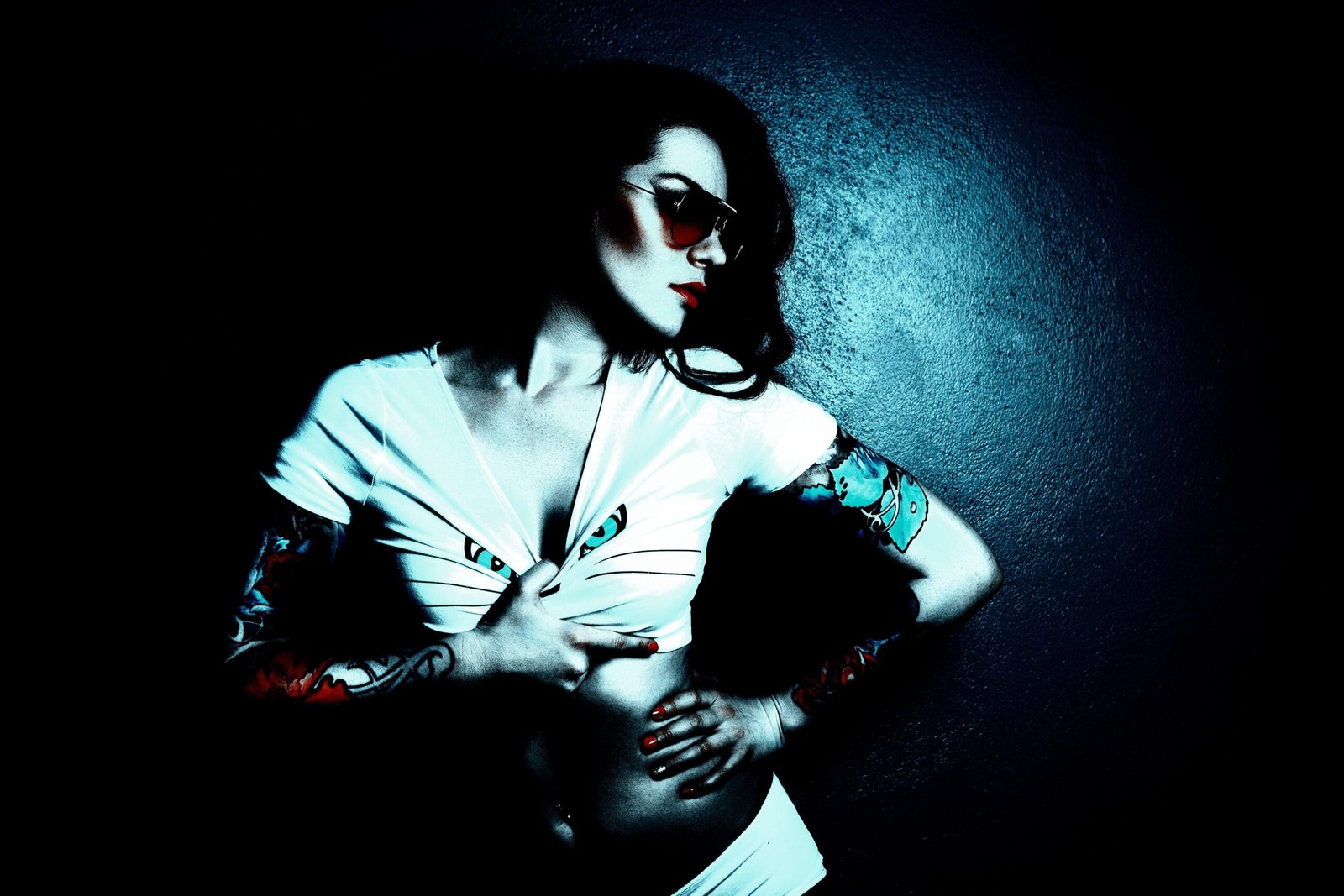 Woman with gothic tattoos on both arms pulling down her white top, with dramatic lighting and dark background.