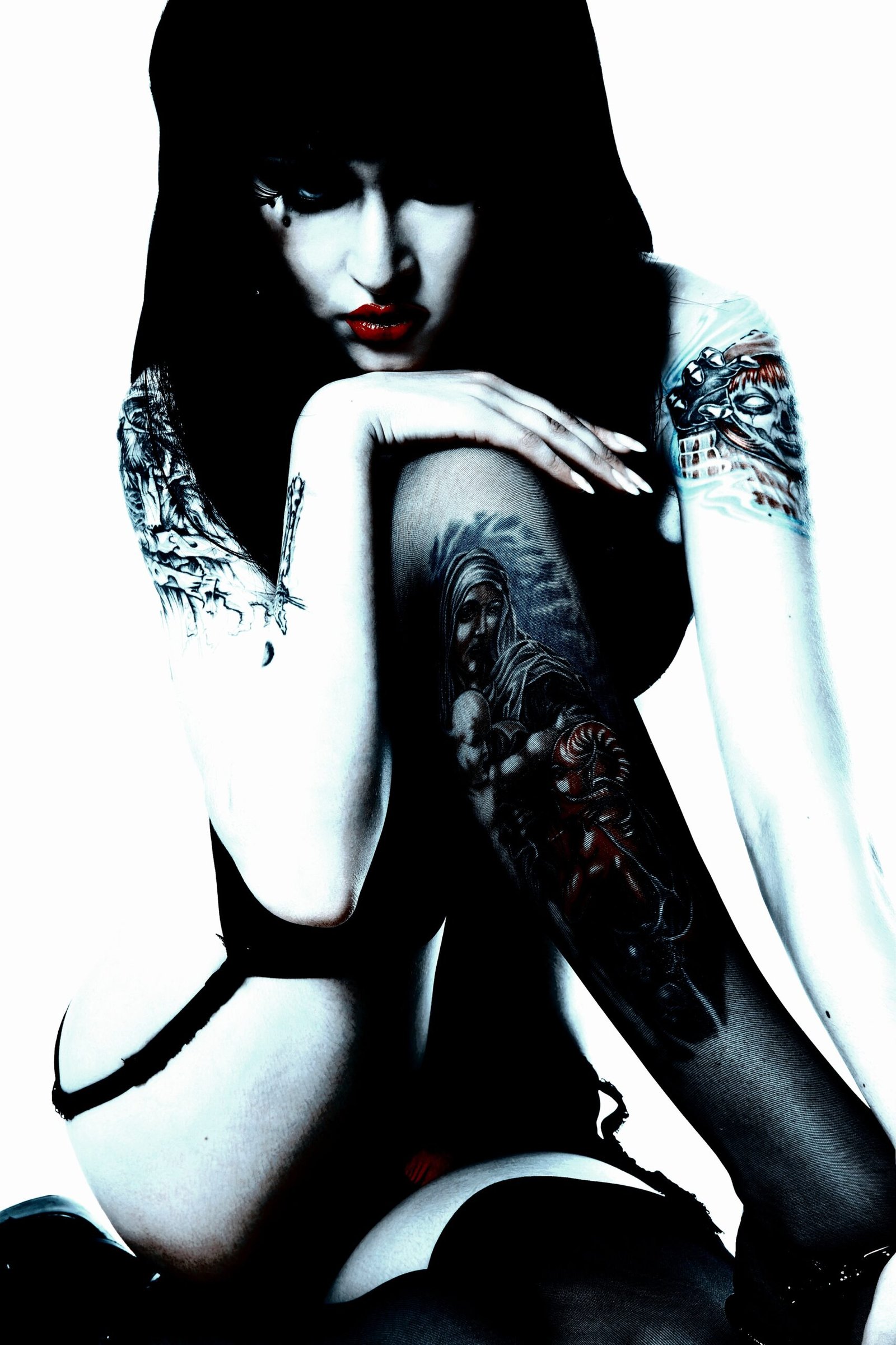 Woman with dark hair and multiple tattoos on her arms and legs, sitting in a contemplative pose with a stark contrast lighting.