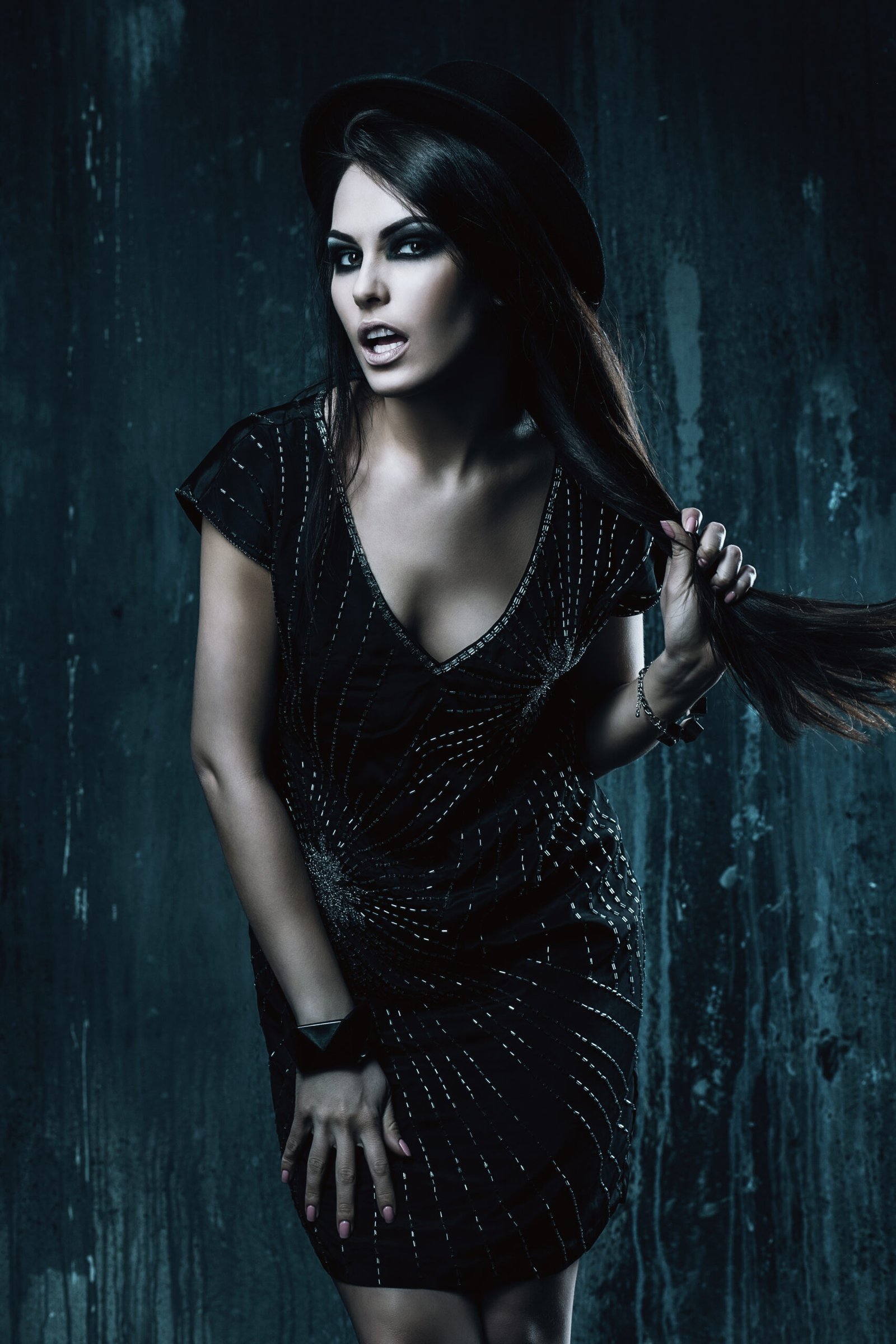 Dark-haired woman in a short, sparkly gothic dress and hat, posing playfully with an intense gaze.