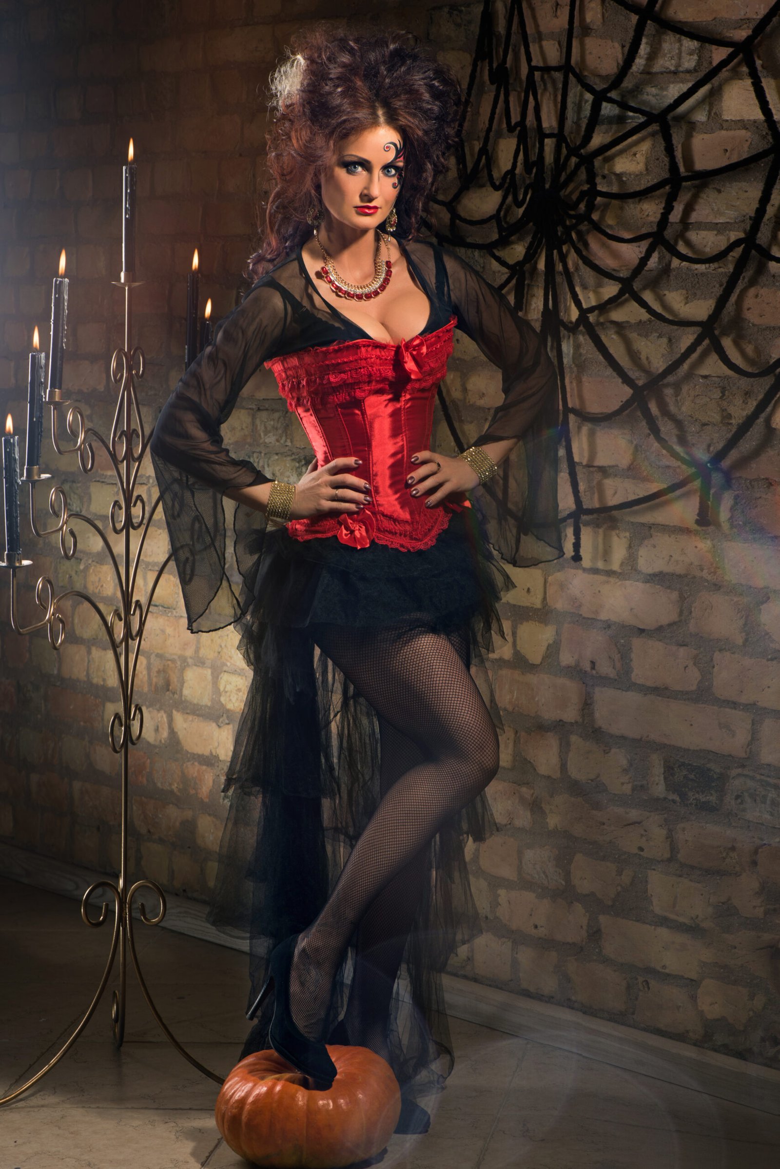 Gothic woman in a red corset dress with black tulle skirt, standing beside a candlelit wall with spider web decor.