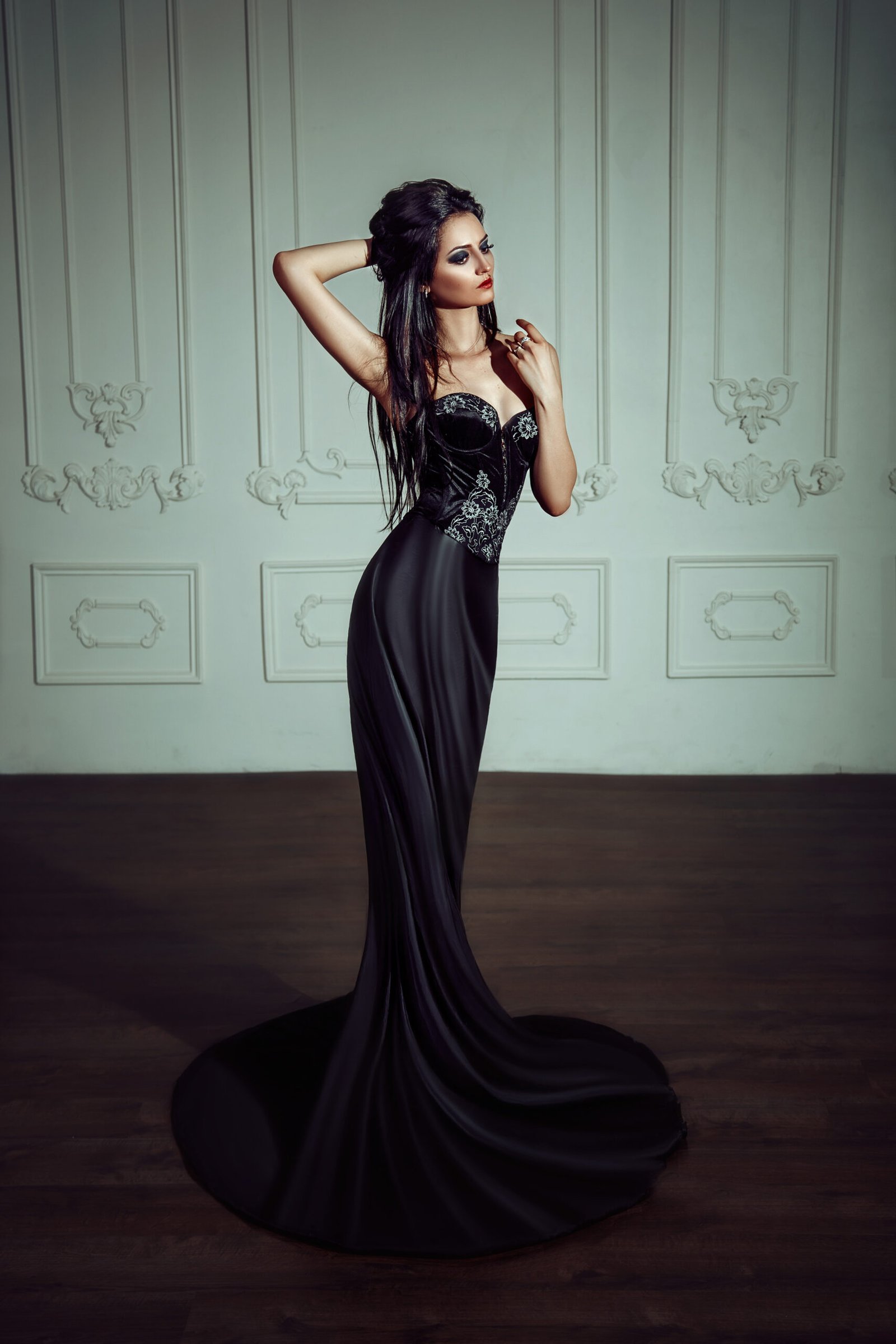 Elegant woman in a gothic black corset gown with intricate silver embroidery, posing in a vintage room.