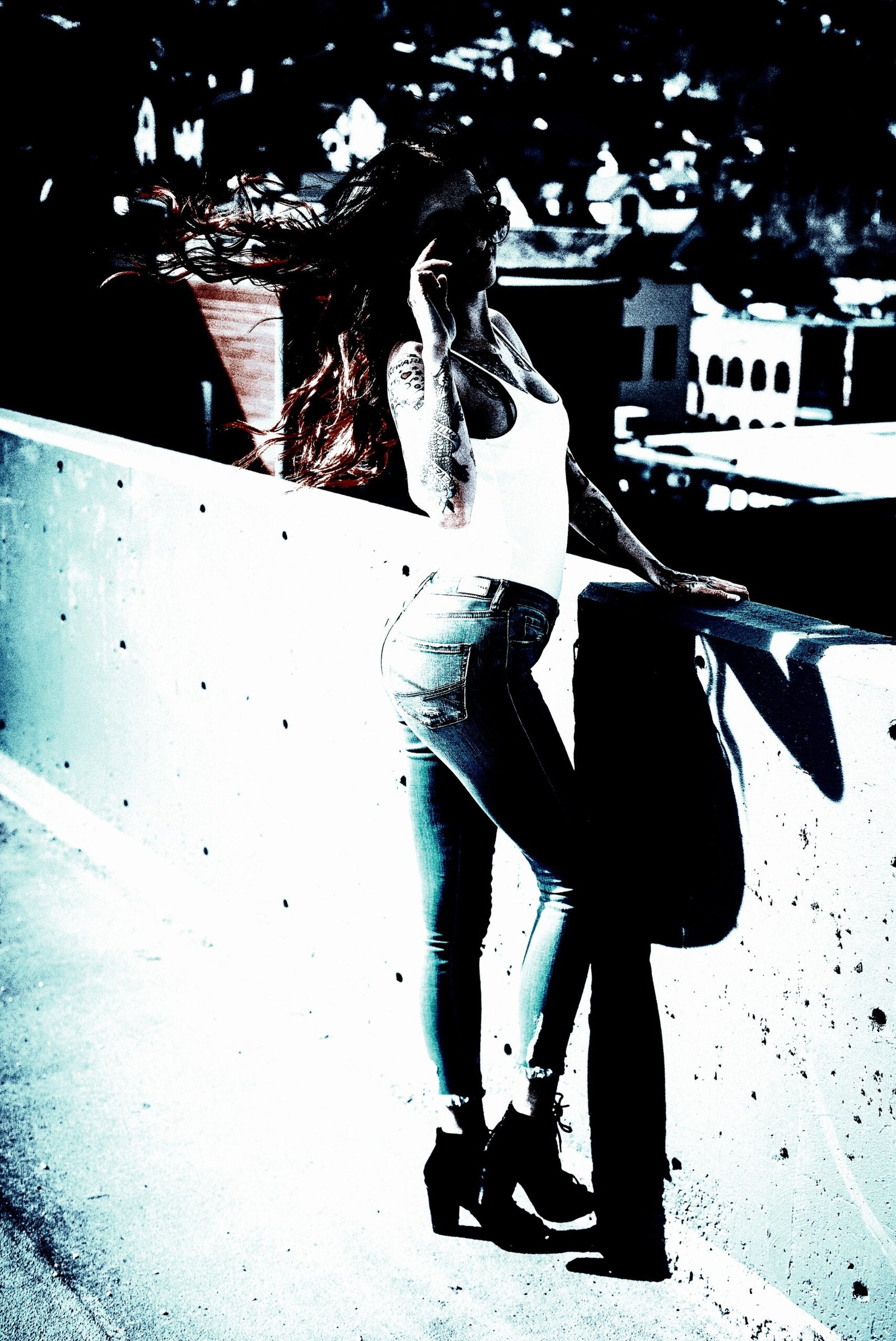 High-contrast image of a woman with long dark hair and red highlights, wearing a white tank top and jeans, posing with aviator sunglasses on a rooftop.
