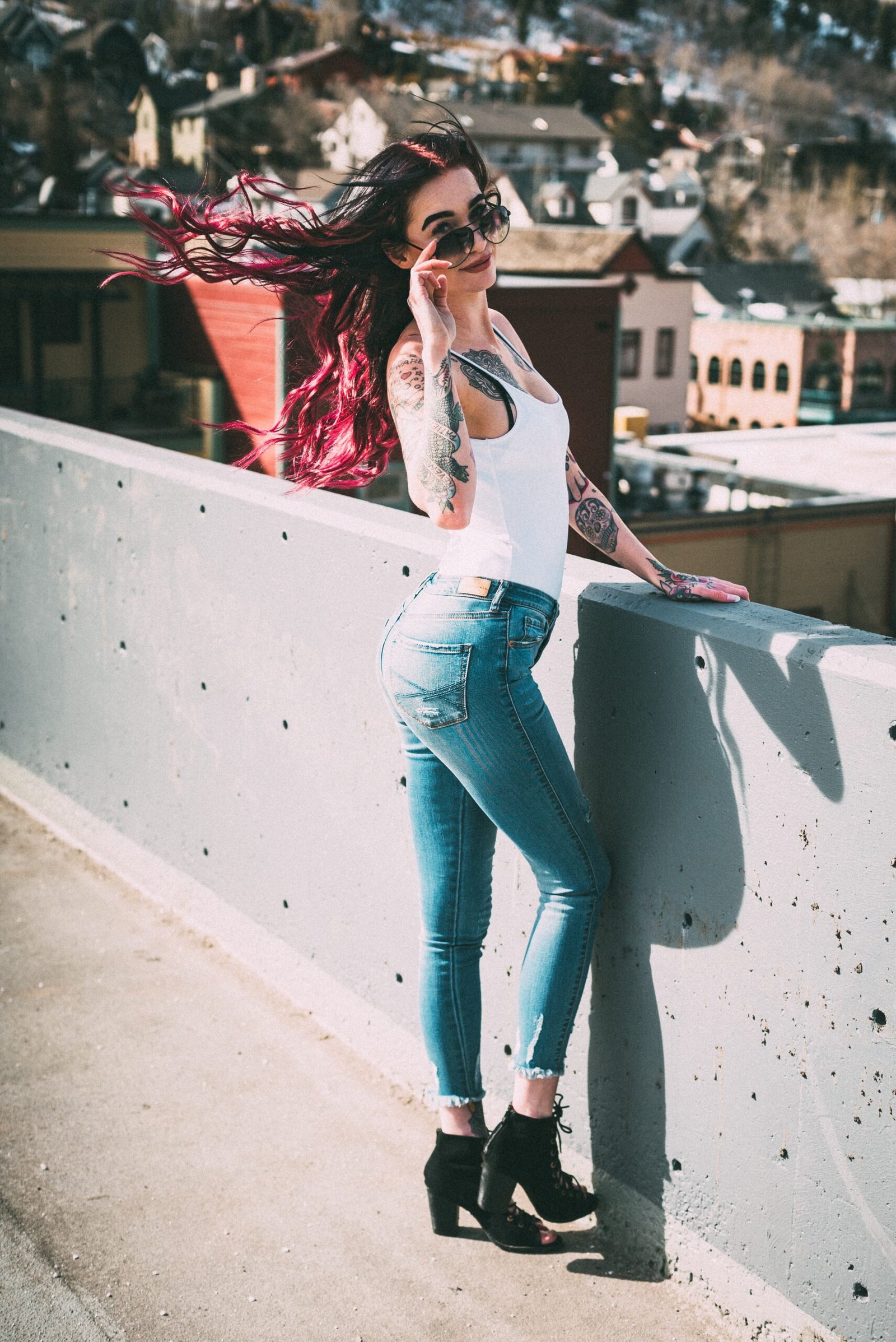 Woman with long dark hair and red highlights, wearing a white tank top and jeans, posing with aviator sunglasses on a rooftop.