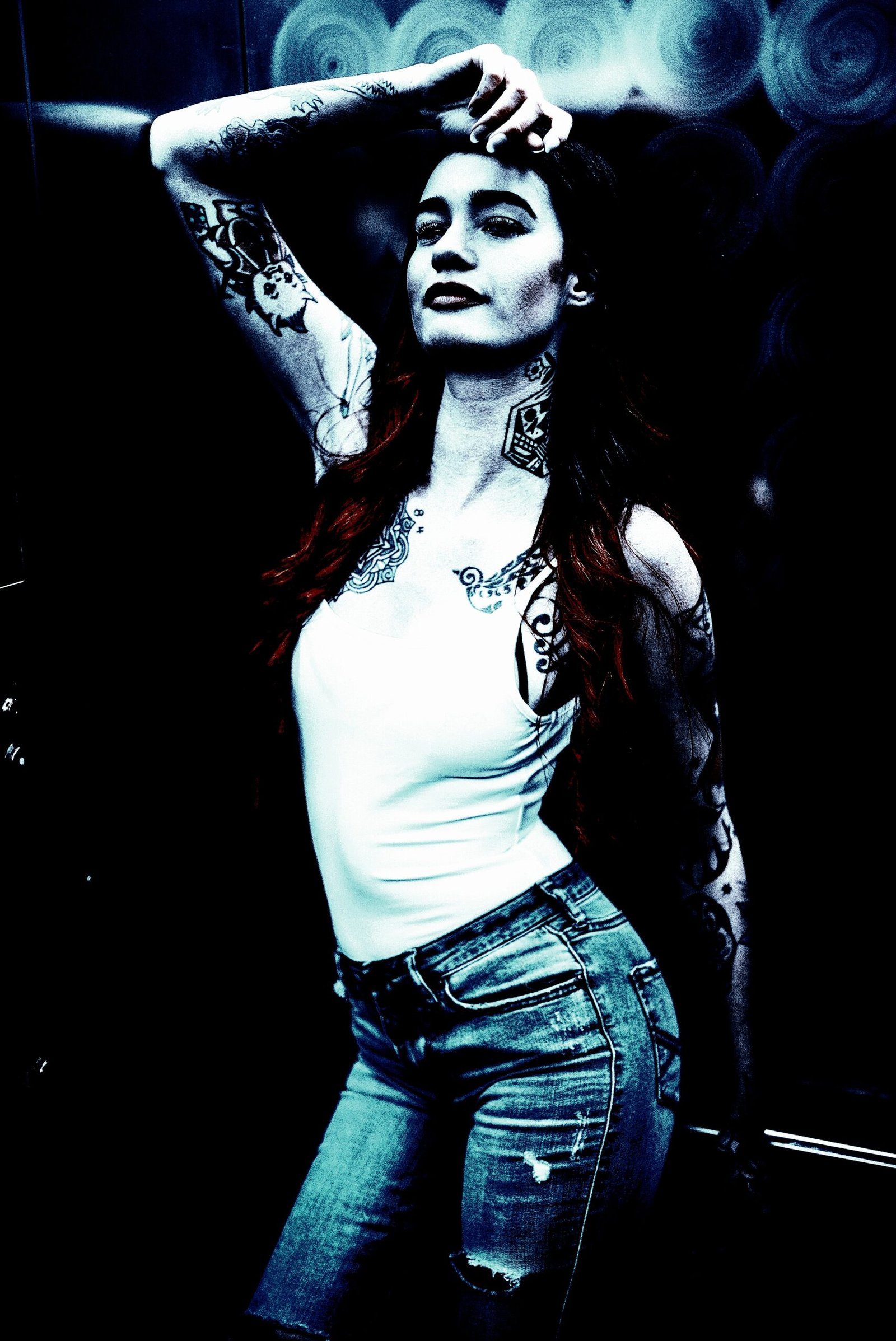 Stylized high-contrast image of a woman with tattoos and long dark hair with red highlights, posing against a metal wall in a white tank top and distressed jeans.