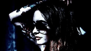 Close-up of a woman with aviator sunglasses, long dark hair with red highlights, and visible arm tattoos, posing in sunlight.
