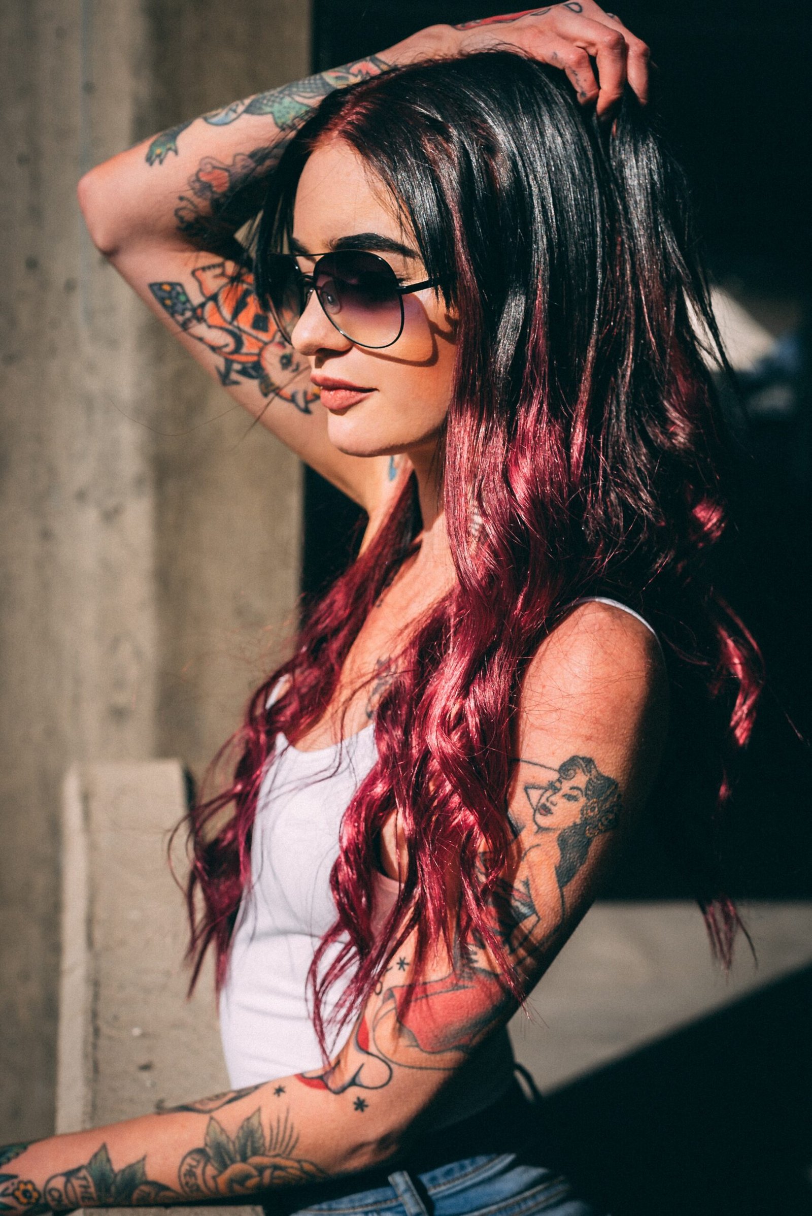 Woman with long dark hair and red highlights, wearing a white tank top and jeans, posing with aviator sunglasses on a rooftop, with hair flowing in the wind.