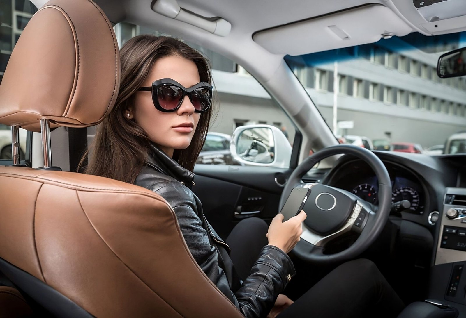 Confident woman in sunglasses and a leather jacket sitting in a luxury car, looking over her shoulder while holding a smartphone.