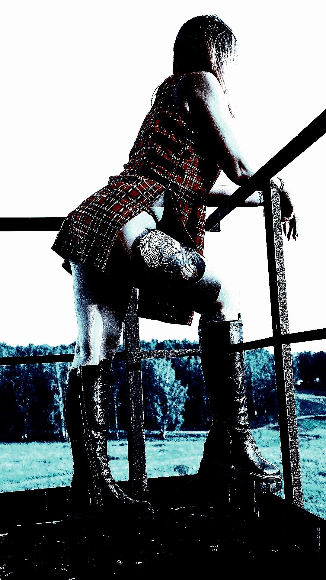 Gothic woman in a red plaid skirt and black combat boots, standing on a metal staircase, overlooking a rural landscape.