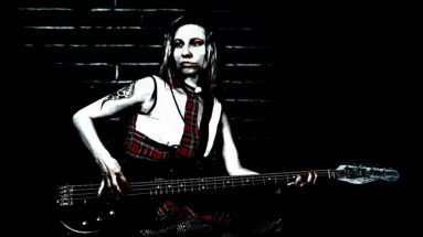 Gothic woman in red plaid outfit playing a bass guitar in a dark, moody setting.