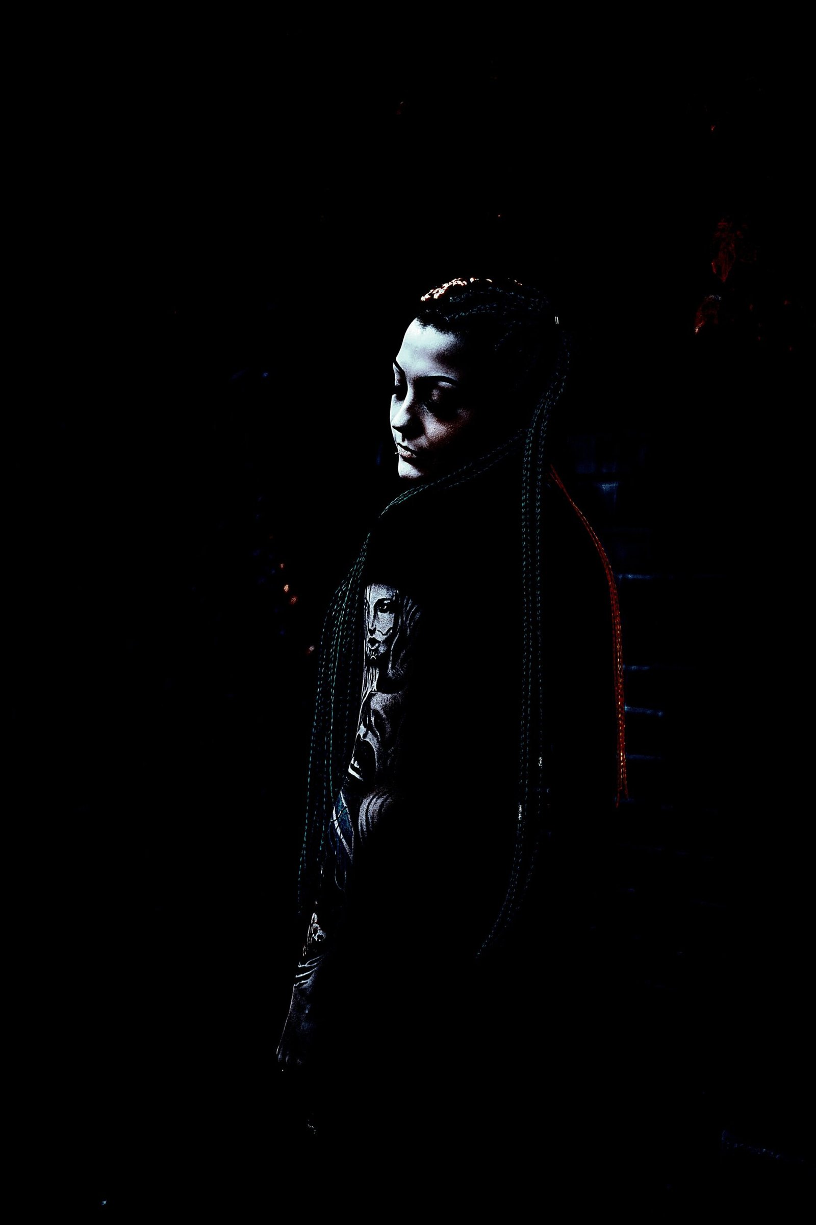 Gothic woman with long braids and intricate tattoos standing in a shadowy environment. 
