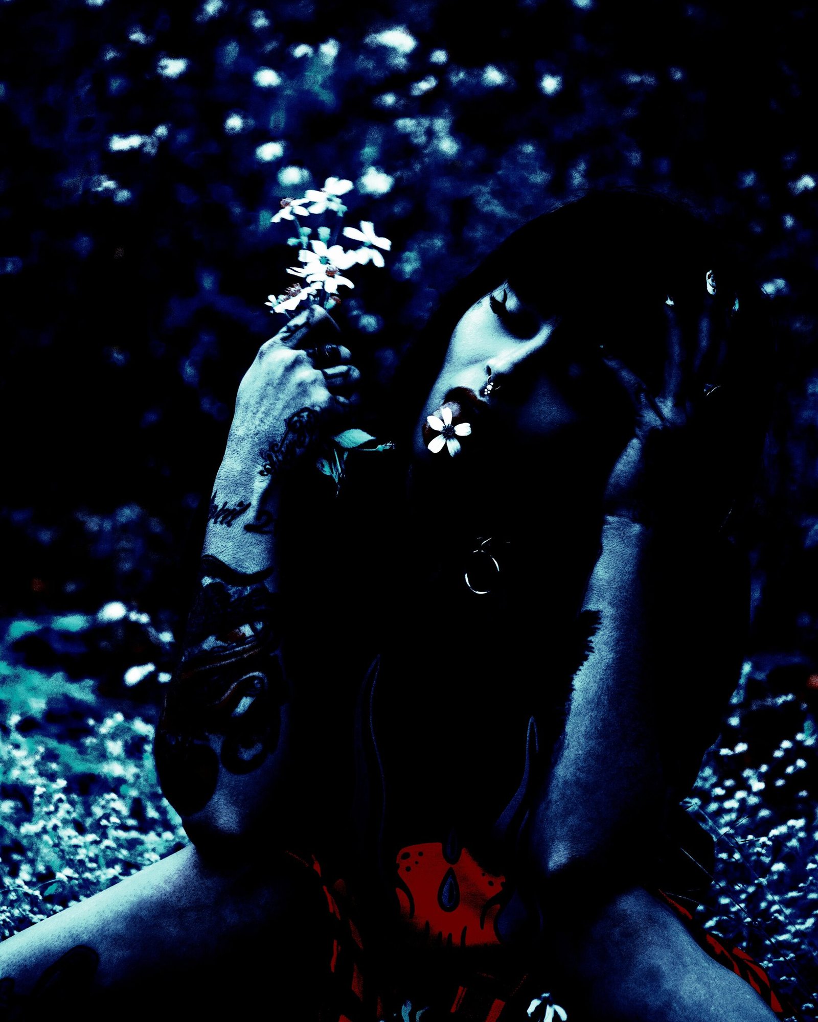 Tattooed woman sitting in a field of flowers, holding a small white flower with a melancholic expression. 