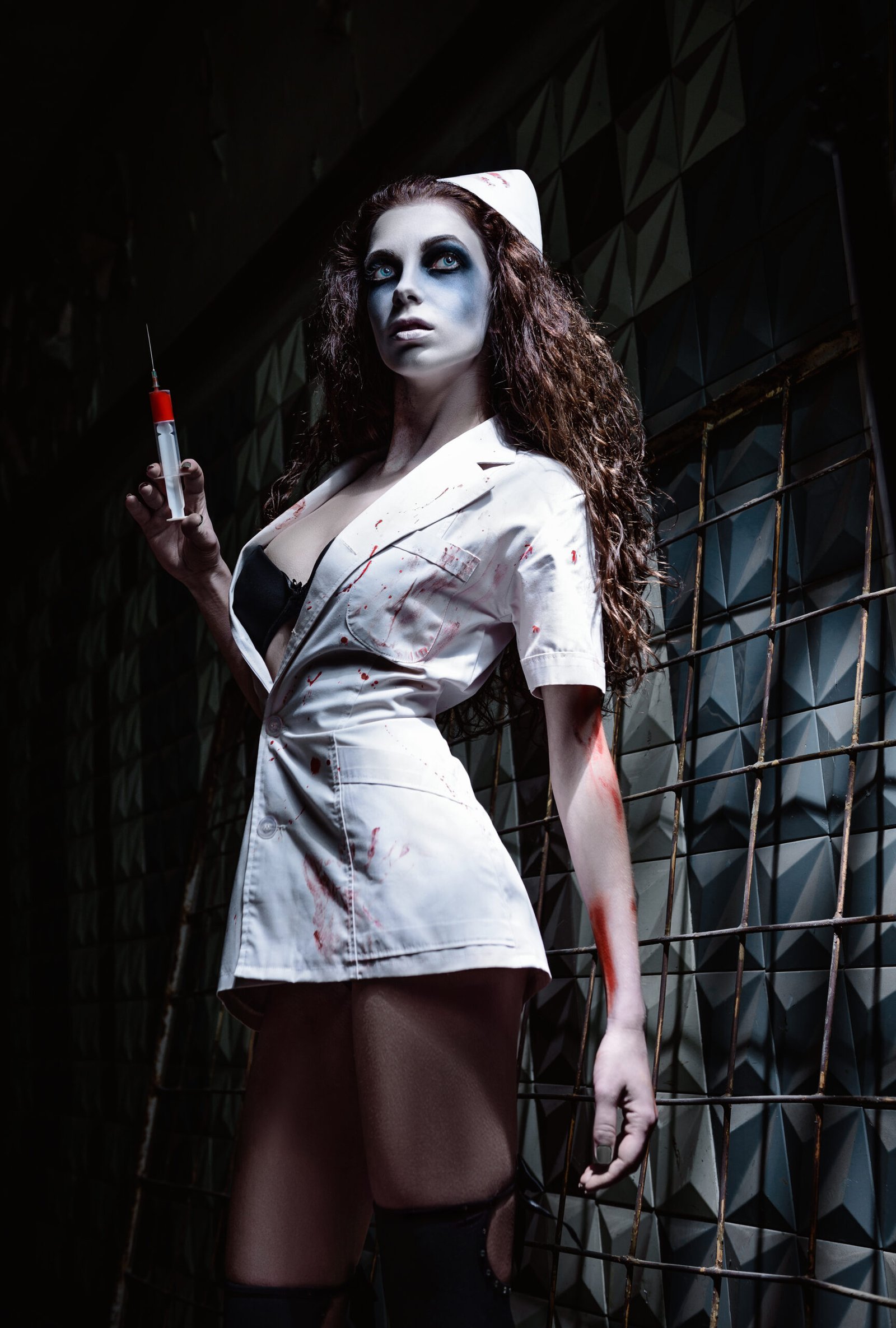 Woman dressed in a blood-spattered nurse costume, holding a syringe in one hand, with dramatic makeup and a dark, eerie setting, portraying a horror-inspired character.