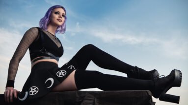 Gothic woman with purple hair in a black mesh top and harness, sitting on a rooftop with a pentagram and crescent moon skirt, showcasing gothic fashion against a sky background.