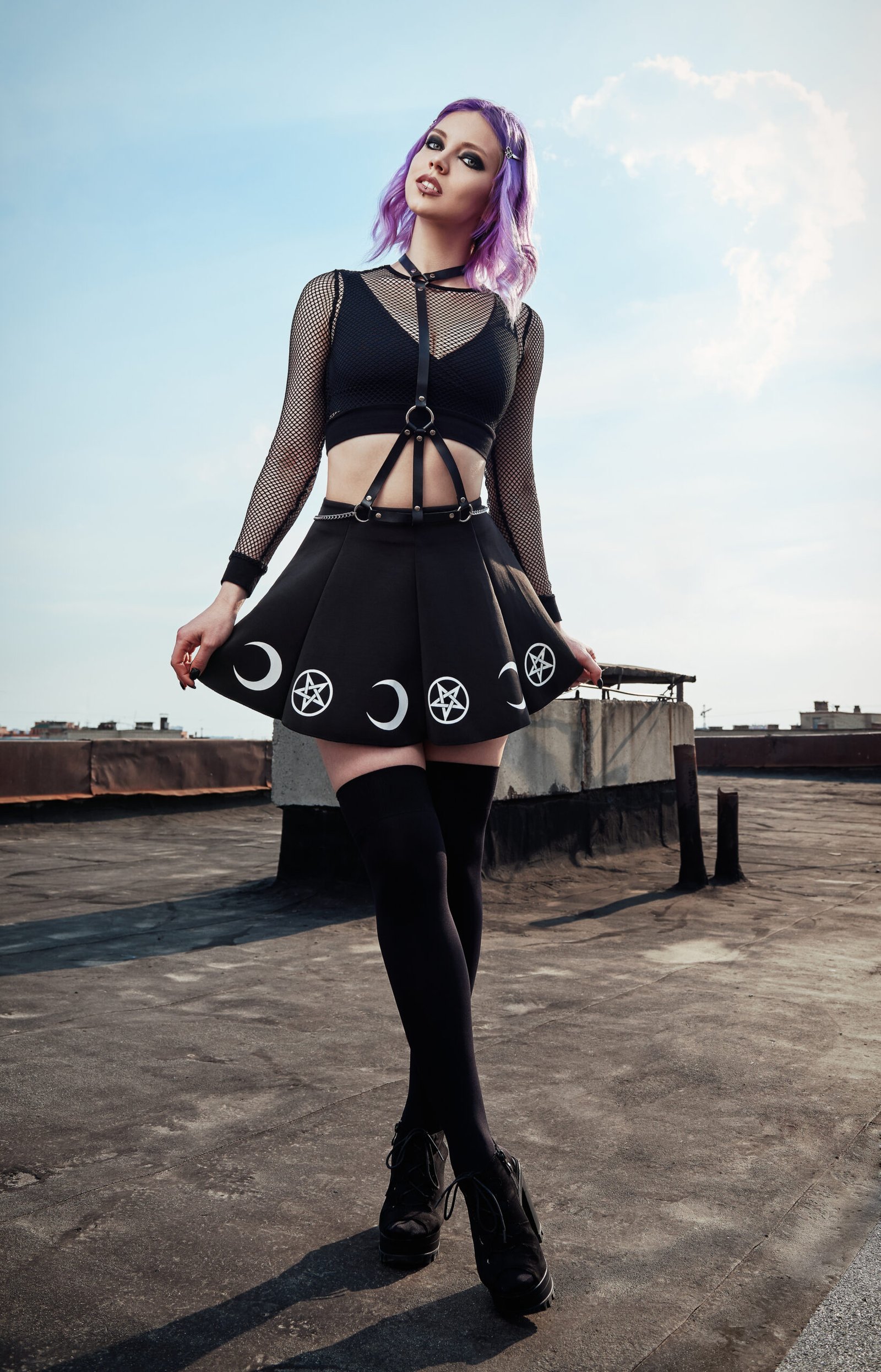 Gothic woman with purple hair standing on a rooftop, wearing a black mesh top, harness, and a pentagram and crescent moon skirt, representing alternative fashion in a bright outdoor setting. 