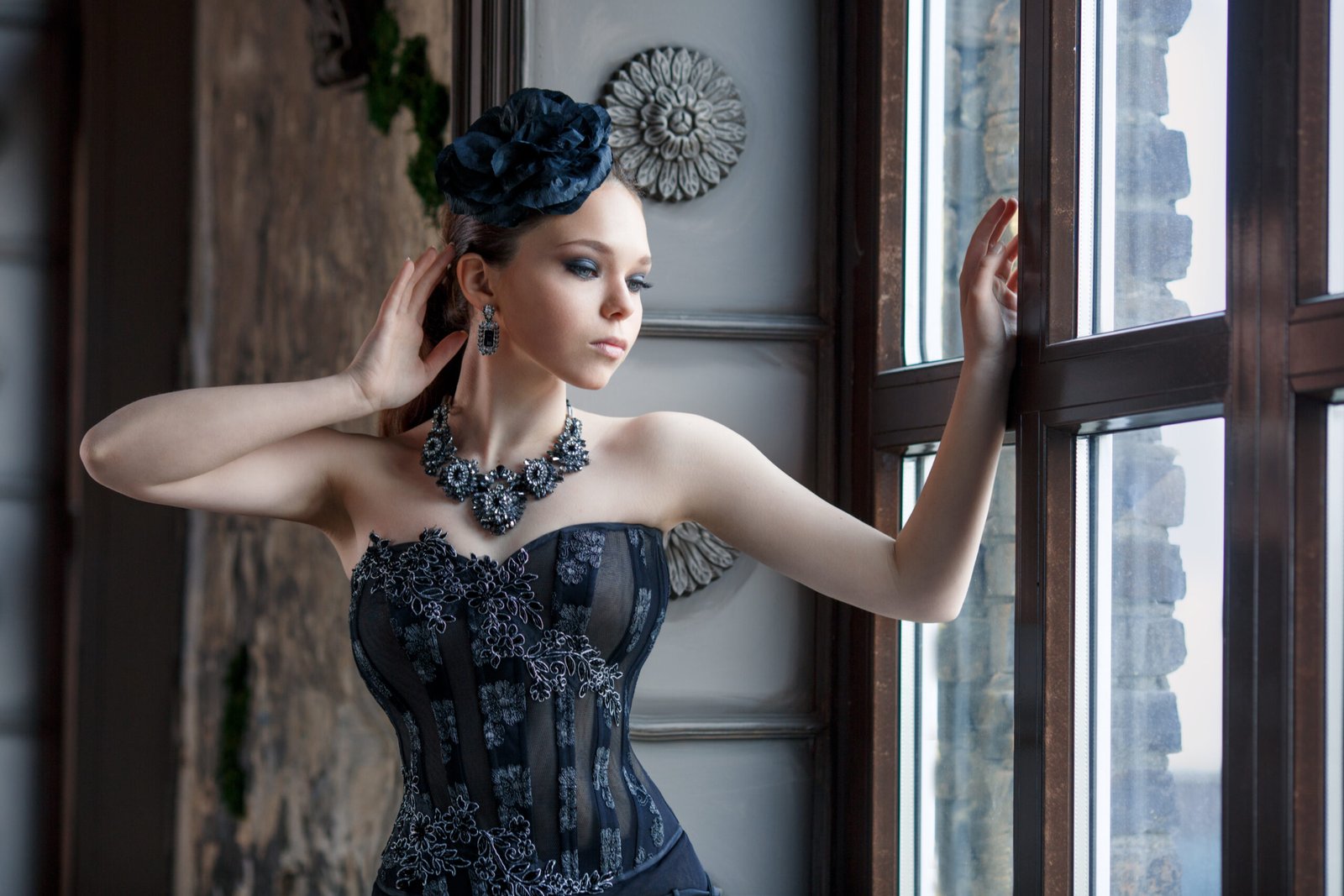 Elegant woman in a Victorian-inspired gothic outfit, featuring a black corset and intricate jewelry, gazing out of a window in a historic setting.