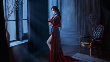 A woman dressed in a long, elegant red velvet gown with lace detailing, standing near a window in a dark gothic room, holding a glass of red wine.