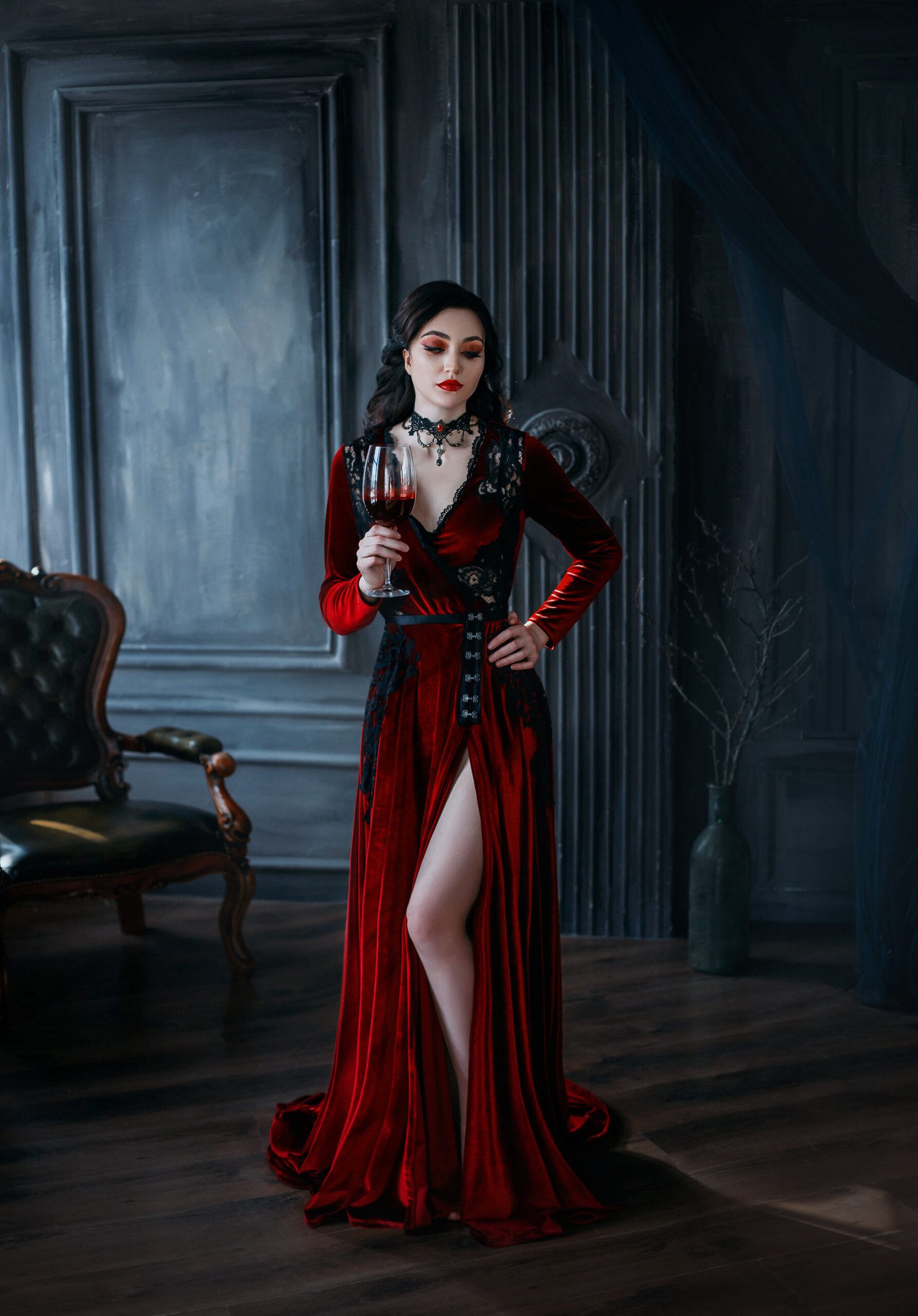 A woman in a full-length red velvet gothic gown with a thigh-high slit, standing in a luxurious dark room, holding a glass of red wine.