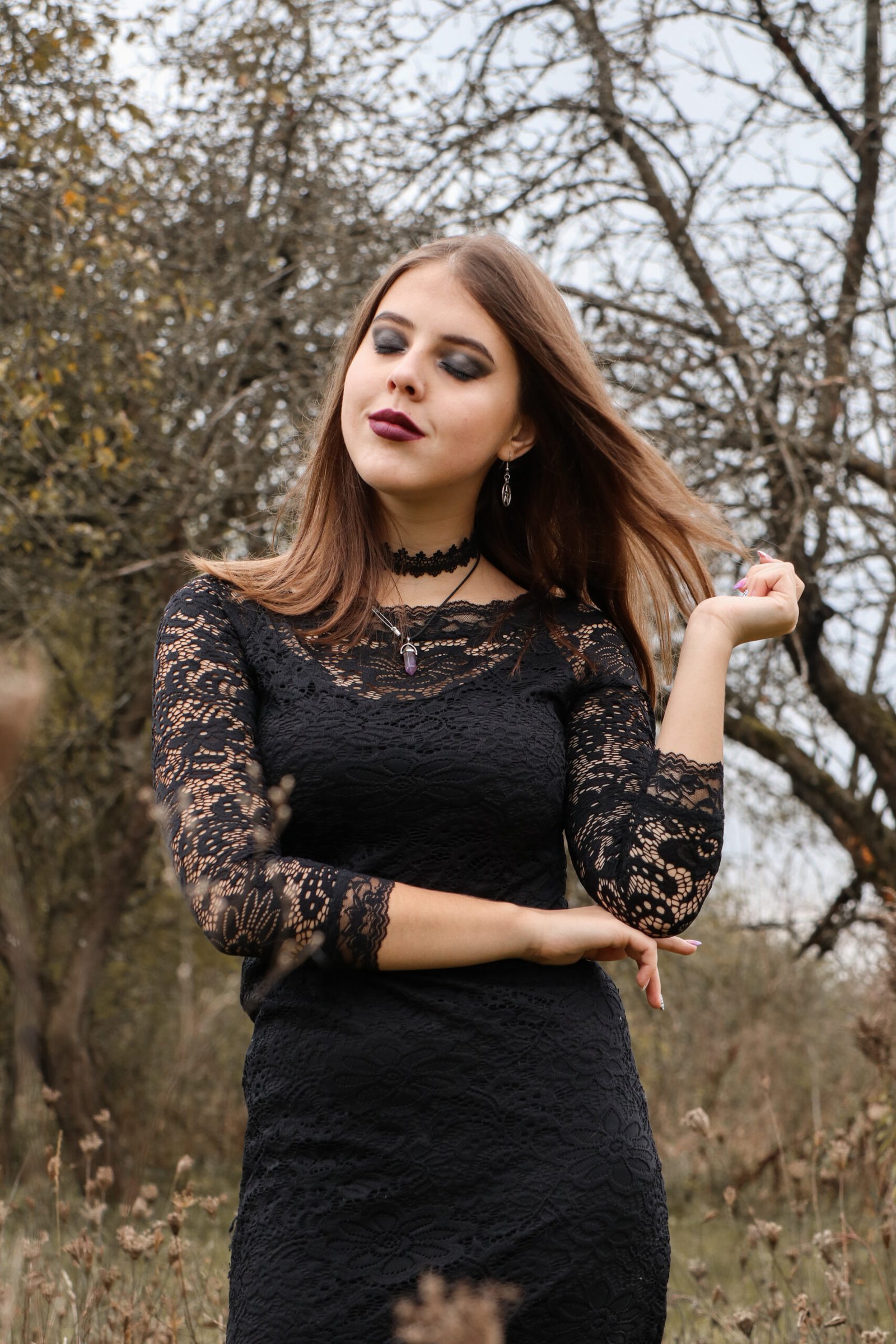 Woman in a black lace dress with a Gothic choker necklace, standing in an autumnal landscape.