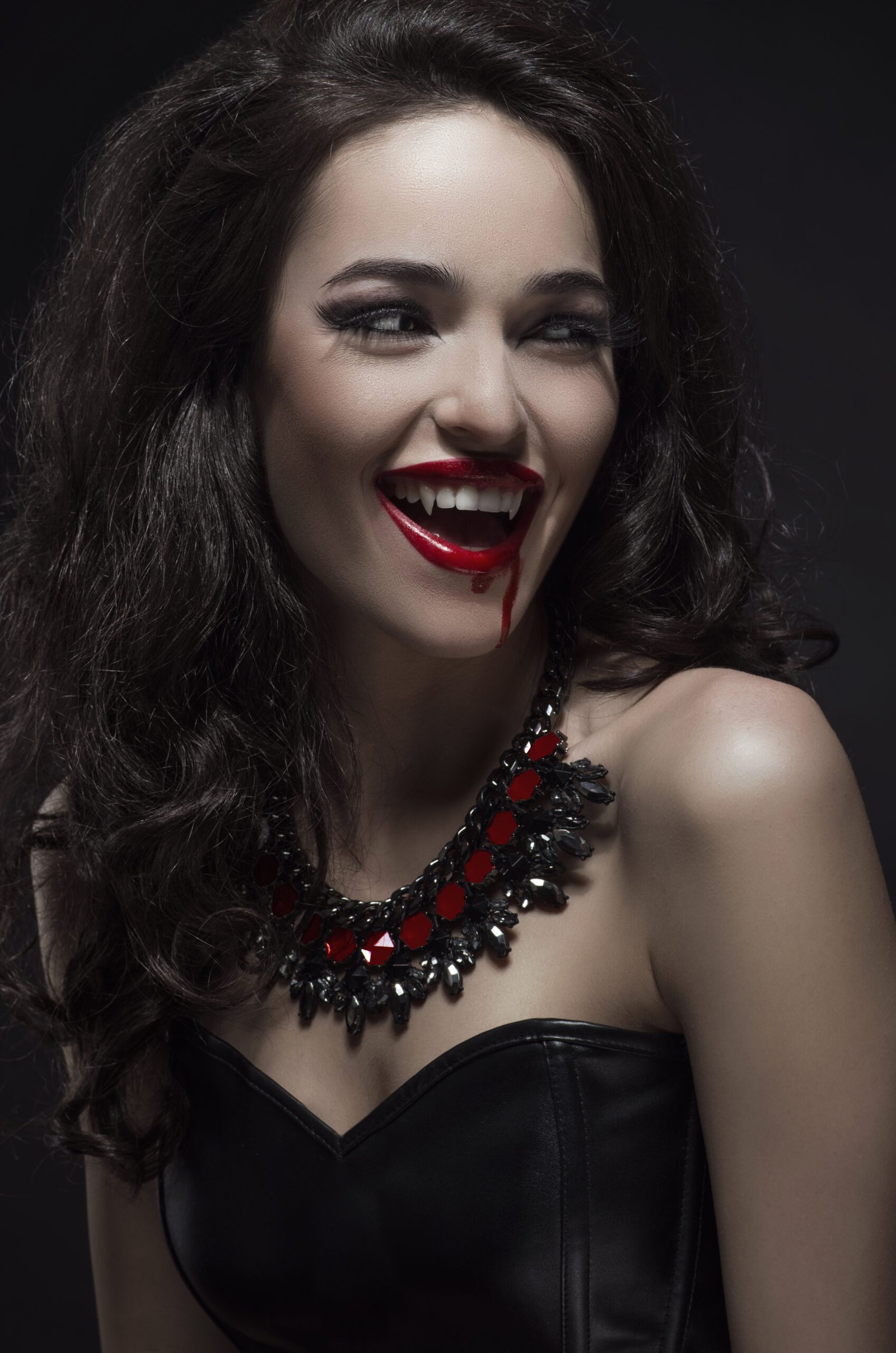 A smiling woman with vampire fangs, dripping blood from her lips, wearing a black leather bustier and a gothic black and red gemstone necklace.