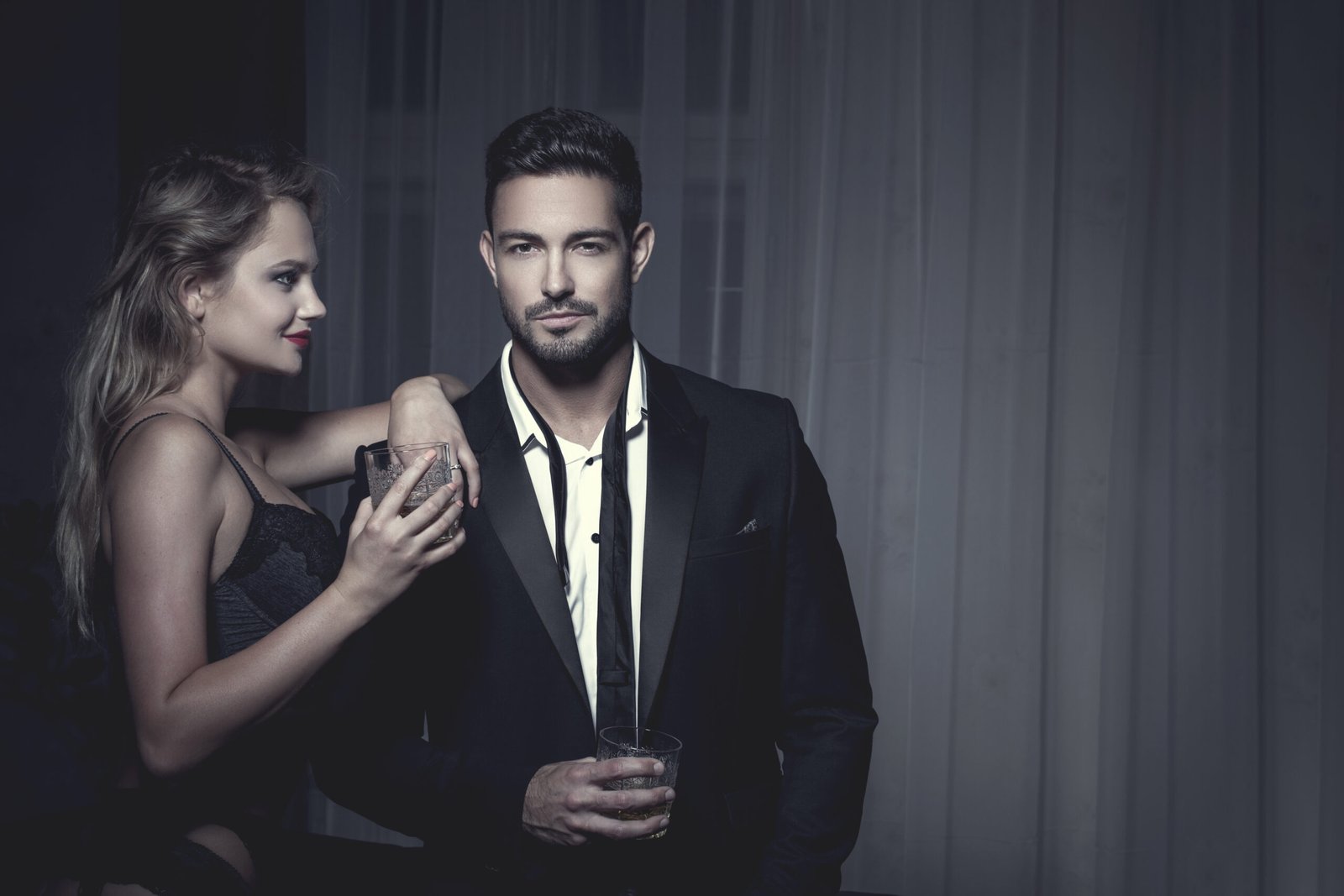 Stylish couple with drinks, the woman leaning on the man's shoulder, both dressed in black formal wear.