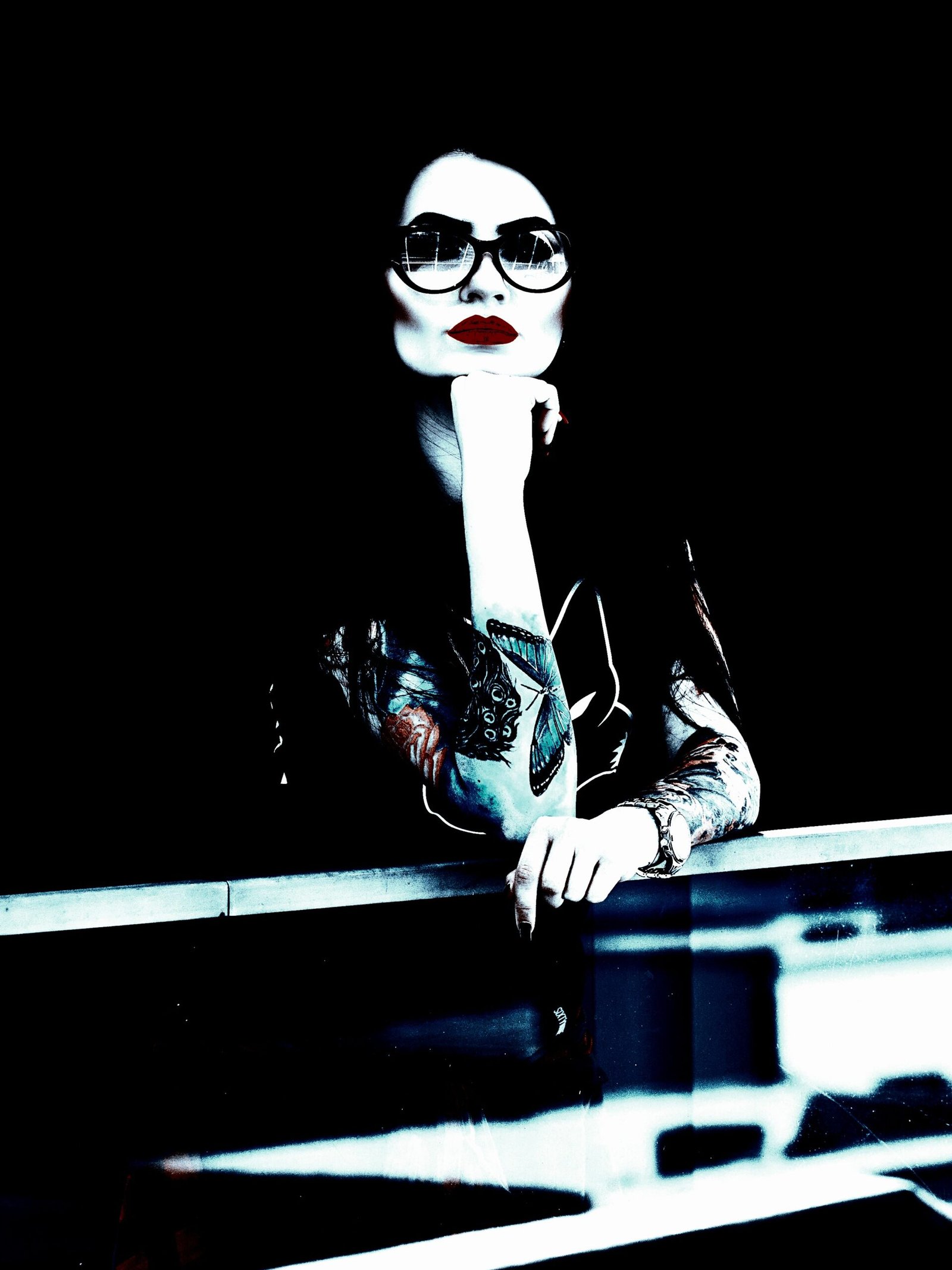 Woman with dark hair and red lipstick wearing sunglasses and a black shirt, leaning on a railing with a contemplative expression.