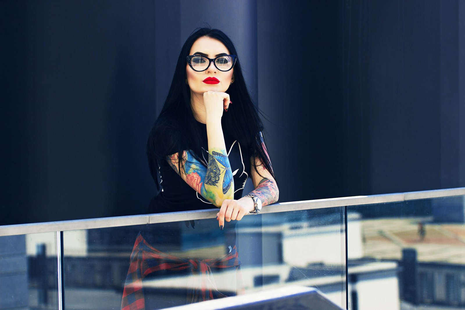 Woman with black hair, wearing glasses, colorful arm tattoos, and a black t-shirt with a fox graphic, leaning on a glass railing in an urban setting, portraying a cool and confident goth aesthetic.
