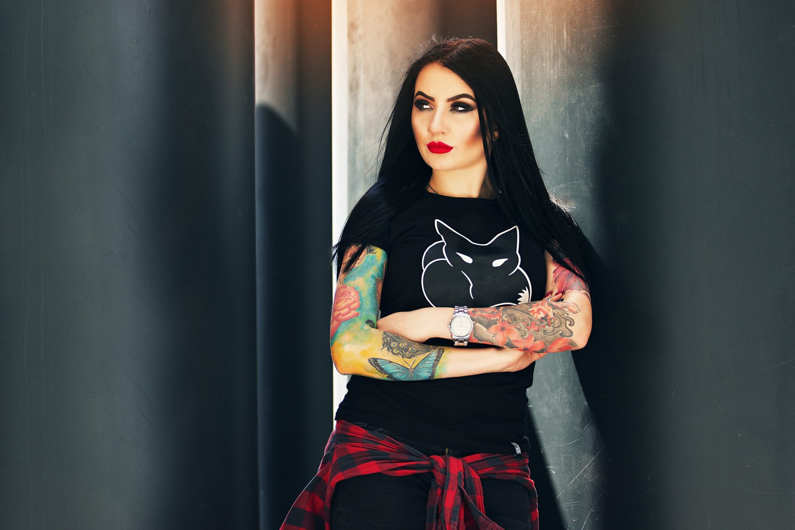 Woman with long black hair, wearing a black t-shirt with a fox graphic, colorful tattoos on both arms, standing with arms crossed and leaning against a dark pillar, exuding a bold, goth-inspired look.