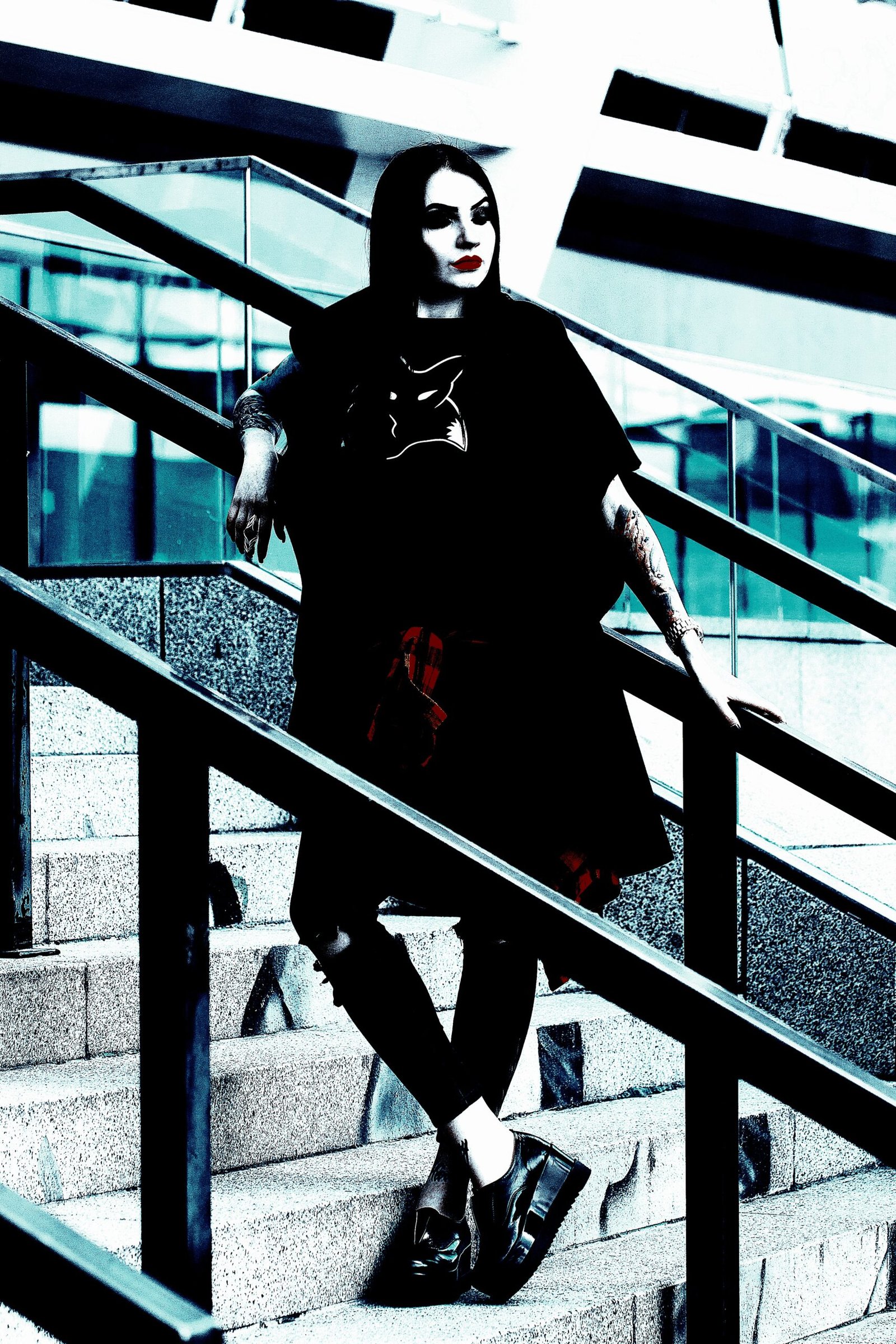 Woman with dark hair wearing a black outfit, leaning on a stair railing, with a confident pose in an urban setting.