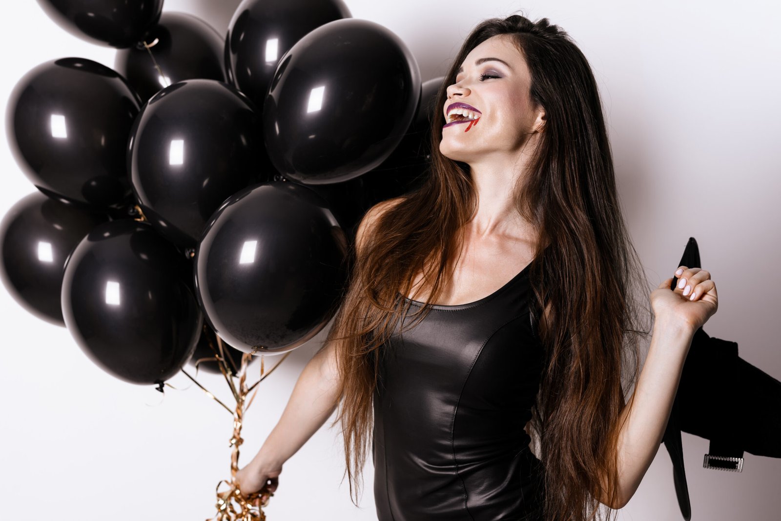 Smiling woman dressed in a black outfit, holding a bunch of black balloons, playfully baring vampire fangs, celebrating with a dark and fun goth aesthetic.
