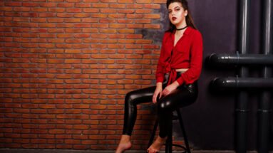 Woman wearing a red shirt and black leather pants, sitting on a stool against a brick wall background.