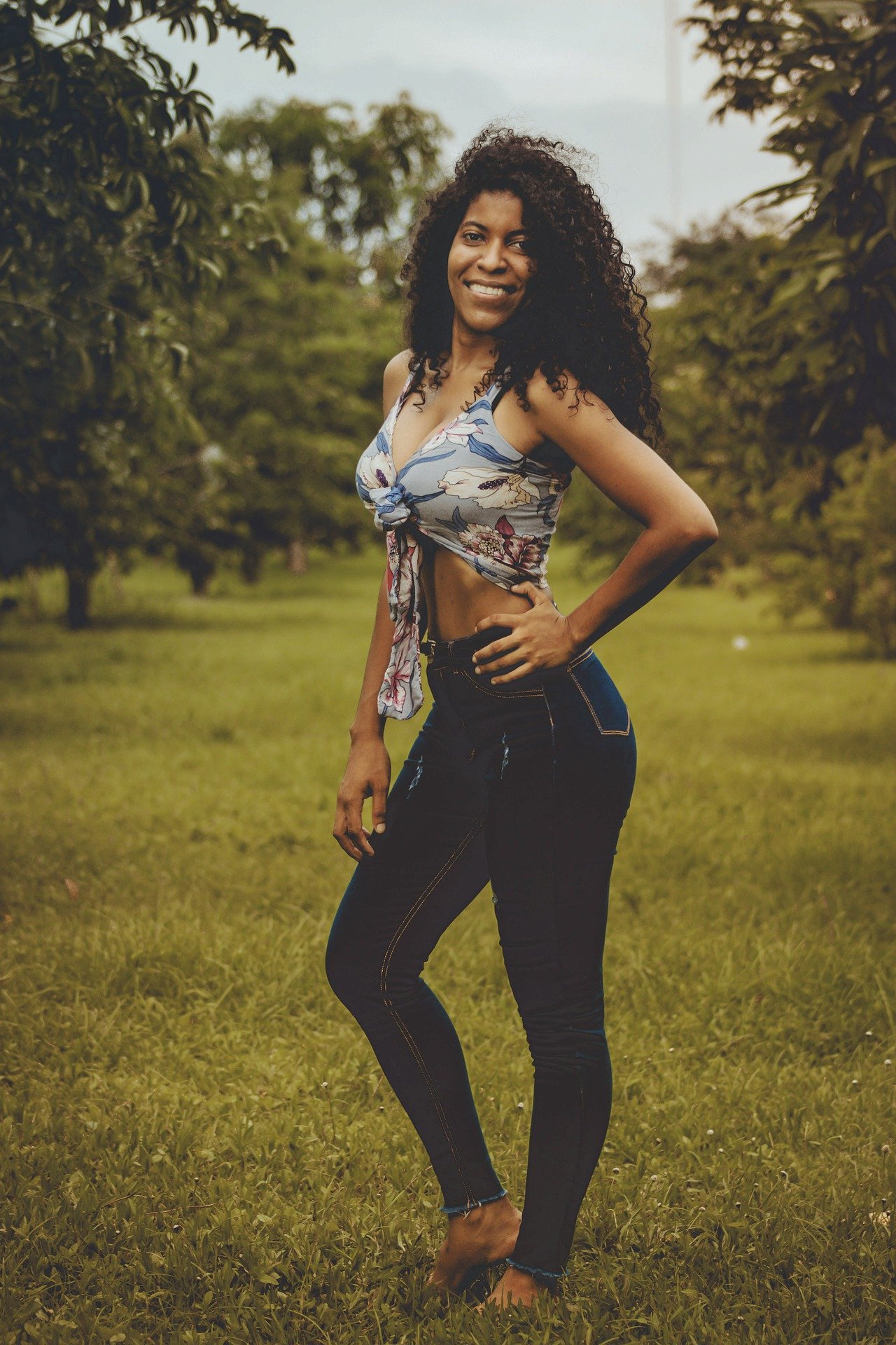  Woman with curly hair smiling, wearing a floral crop top and jeans, standing barefoot in a grassy field.