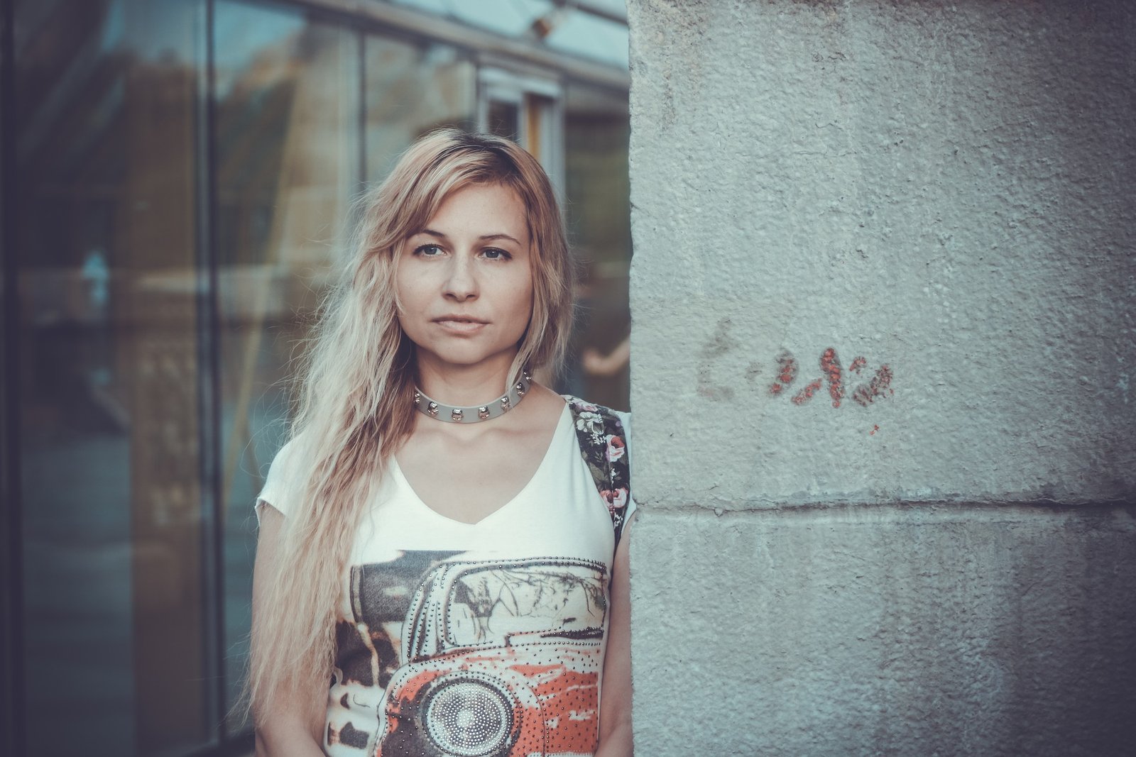 woman with blonde hair wearing a vintage t-shirt and spiked choker, standing by a wall with a calm expression. 